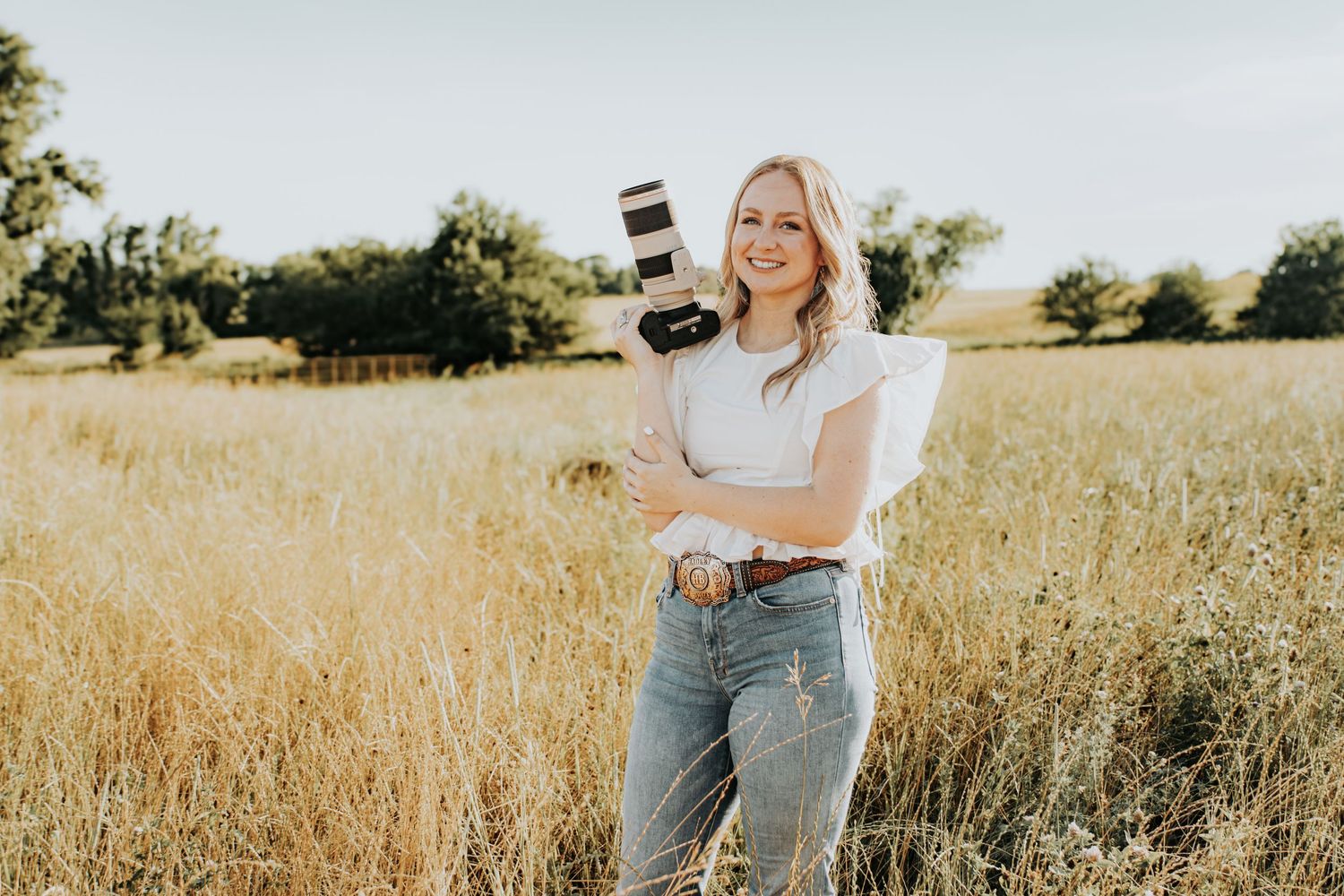 Photographer stands in golden field at sunset holding professional camera while wearing casual white top and jeans.