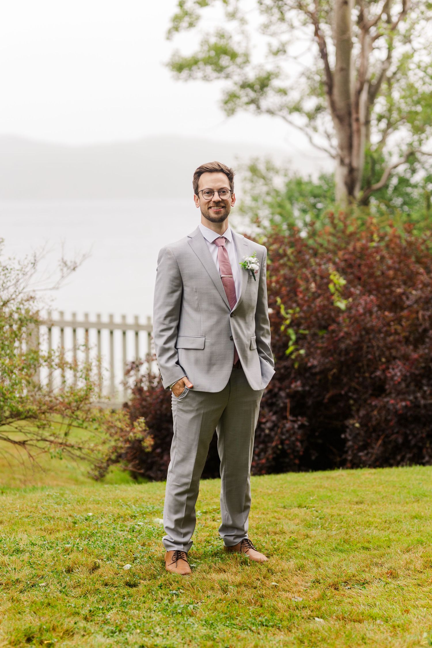 A person in a light gray suit and red tie poses playfully on a grassy hillside with white fence and trees in background.