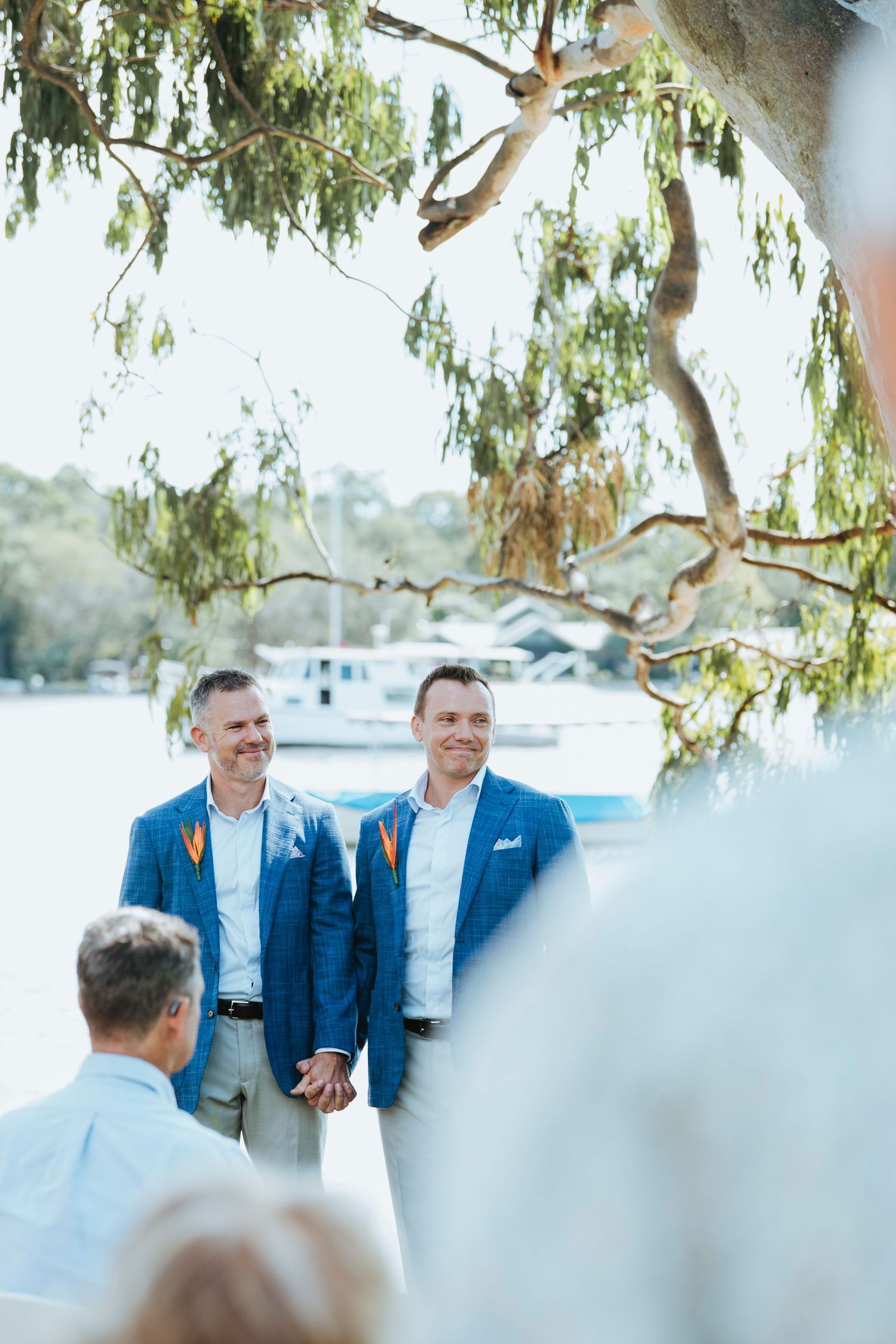 Two men in navy suits share a candid moment under hanging tree branches near a waterfront setting.