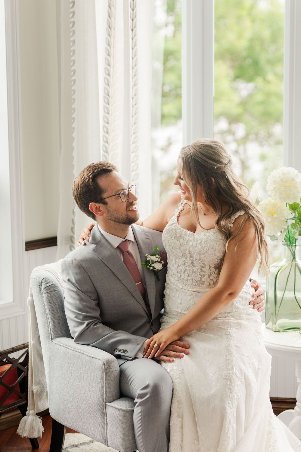 Newlywed couple shares joyful laughs while sitting together on a white armchair in a bright sunlit room.