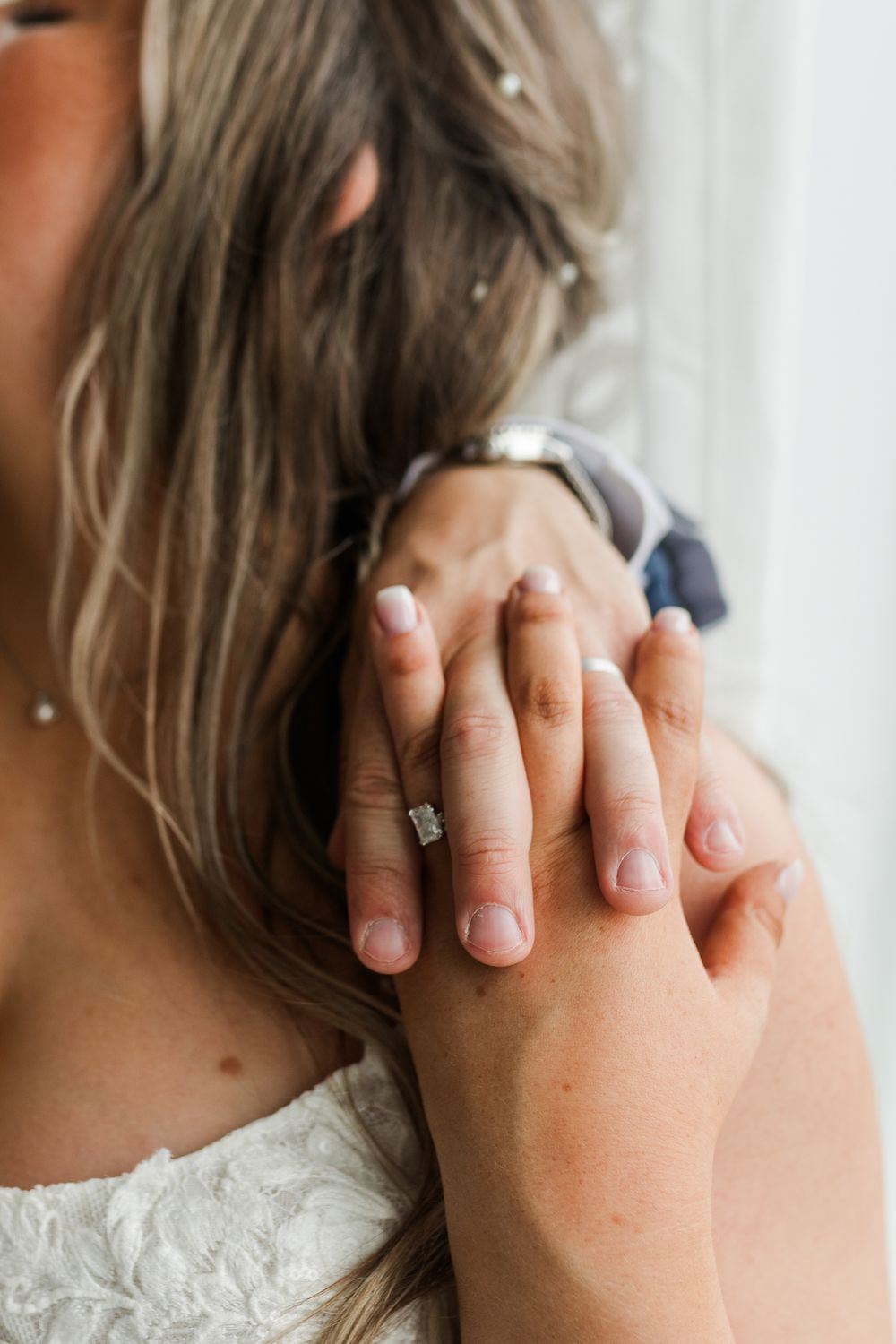 Close-up of hands with engagement ring showing delicate diamond solitaire against white lace dress fabric.