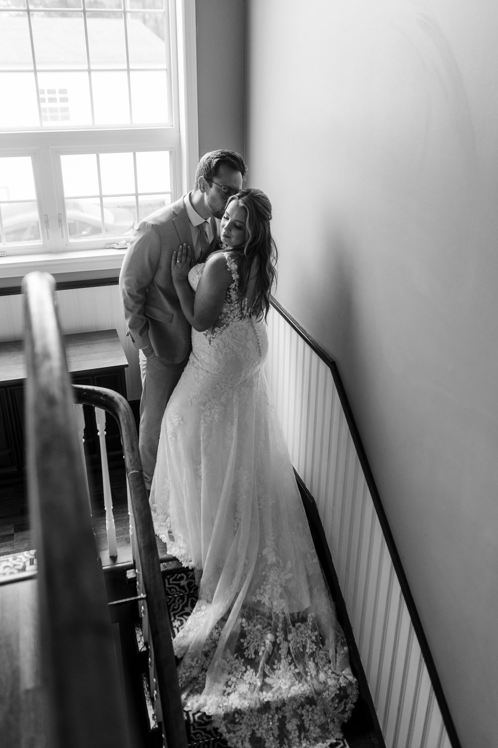 Black and white photograph of a romantic couple in wedding attire standing by a sunlit window on stairs.