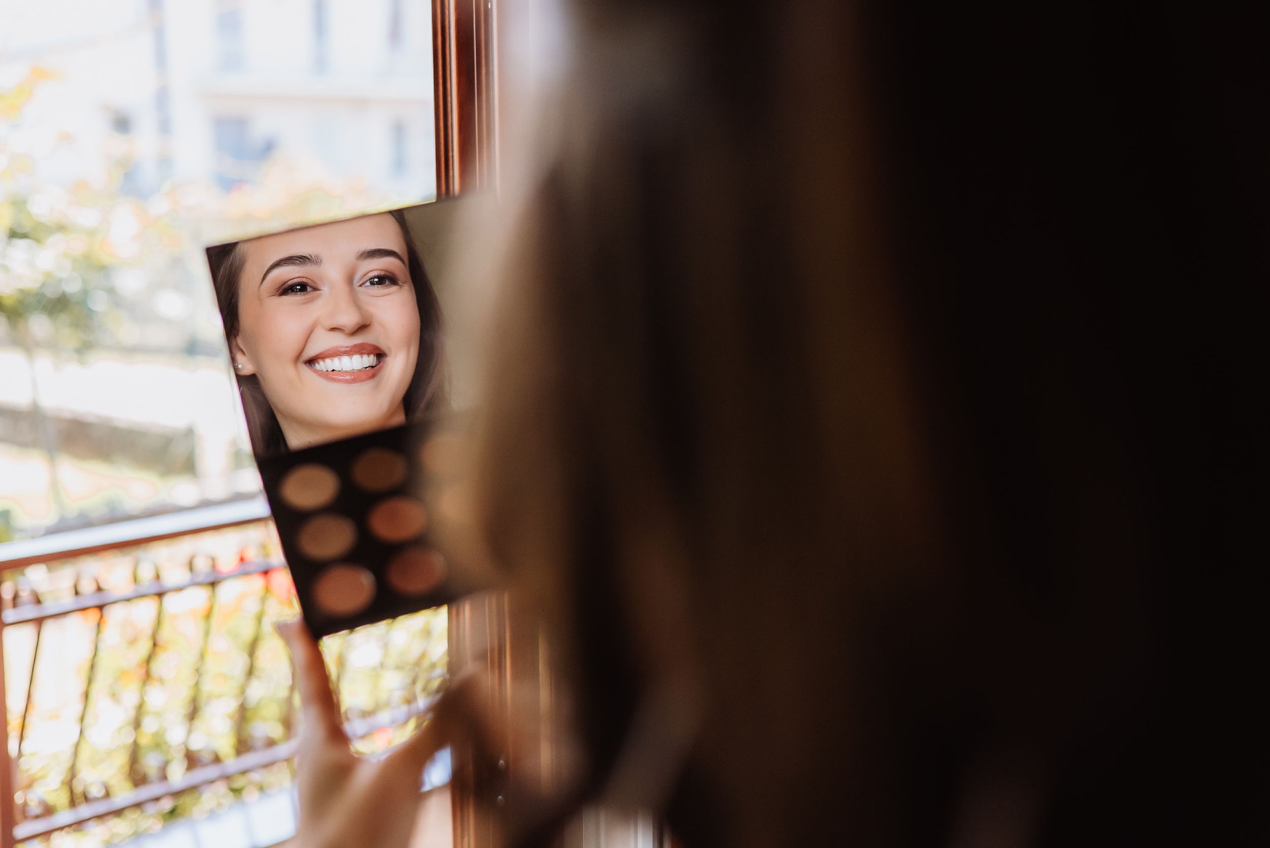 Woman smiling while looking at laptop outdoors on balcony with sunlit background.