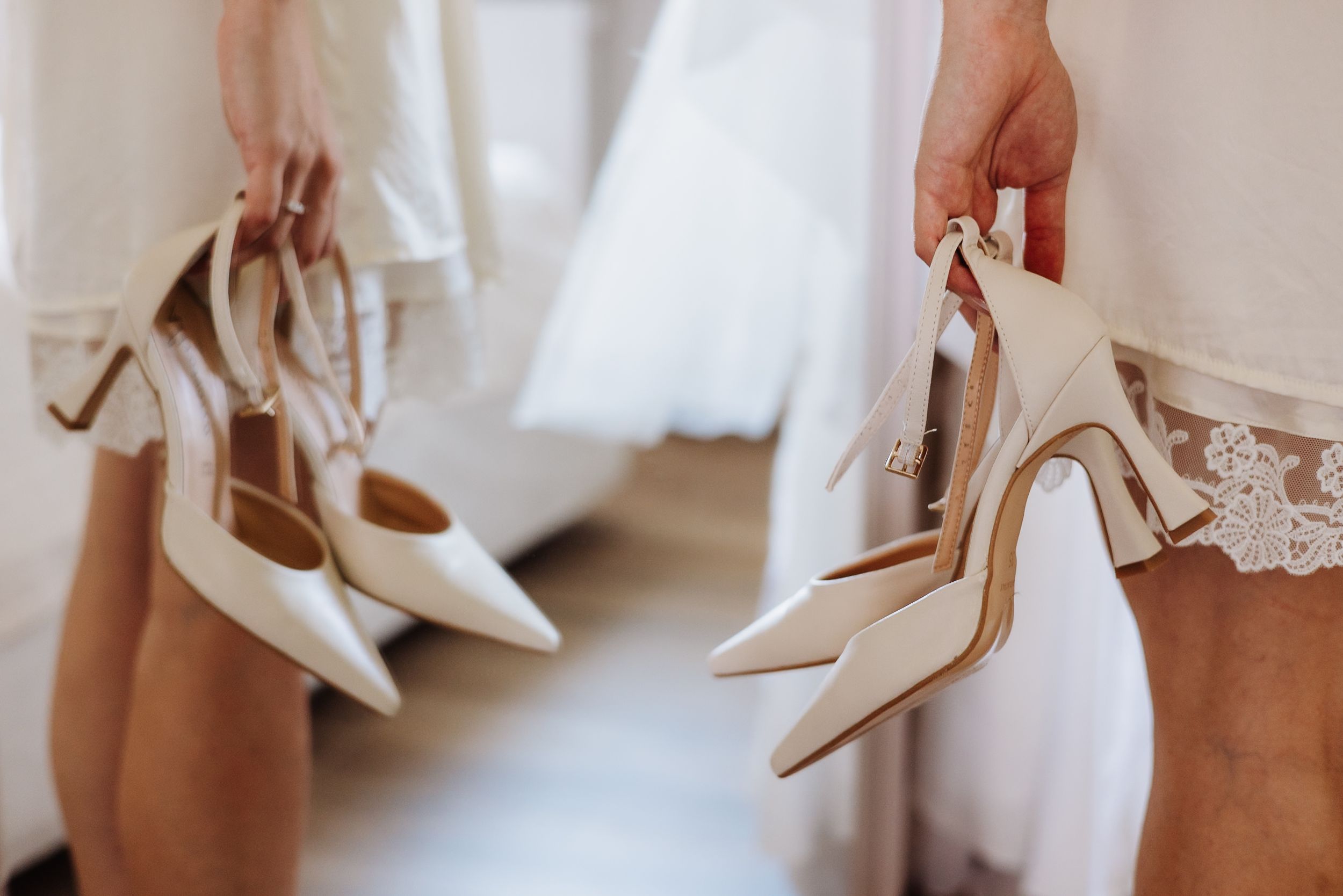 Delicate cream colored high heeled wedding shoes arranged on white fabric with intricate lace details.