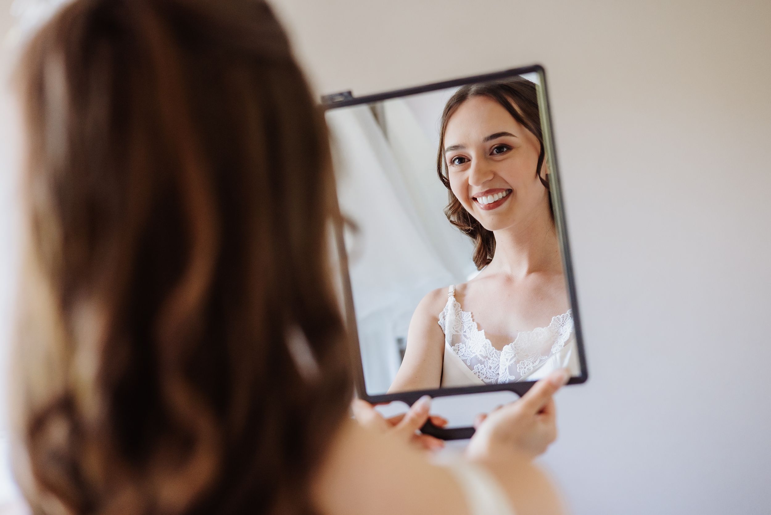 A bride looks at her reflection in two mirrors while having her hair and makeup done for her wedding day.