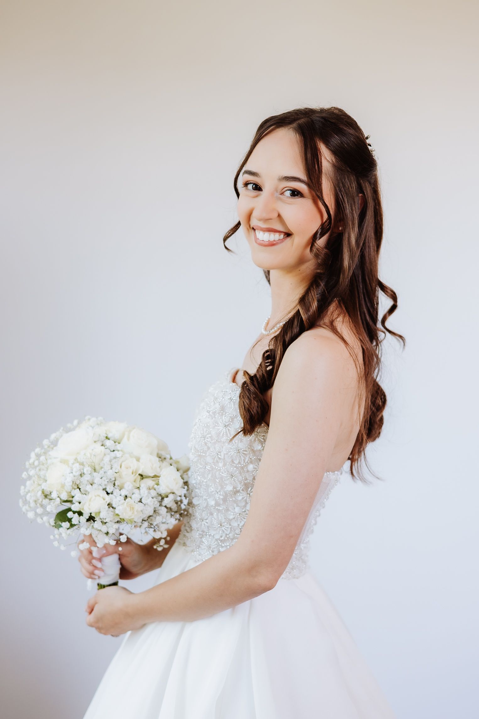 Bride in classic white dress holds a bouquet of white hydrangeas while posing against a soft light background.