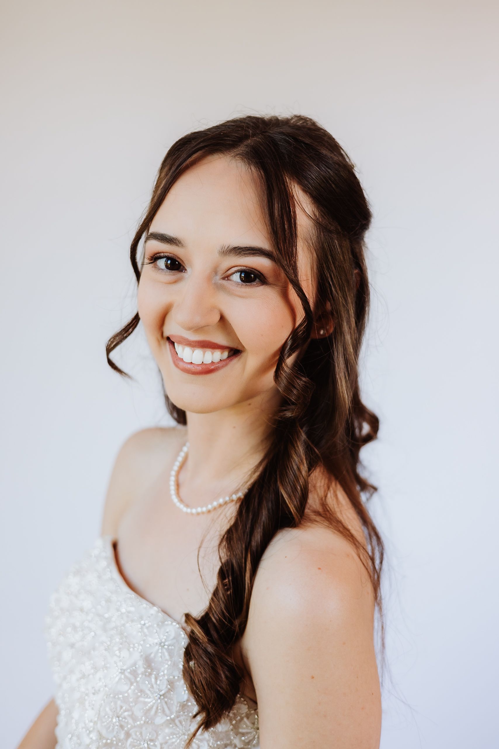 A joyful bride wearing a white lace wedding dress and pearl necklace smiles brightly in a bridal portrait series.