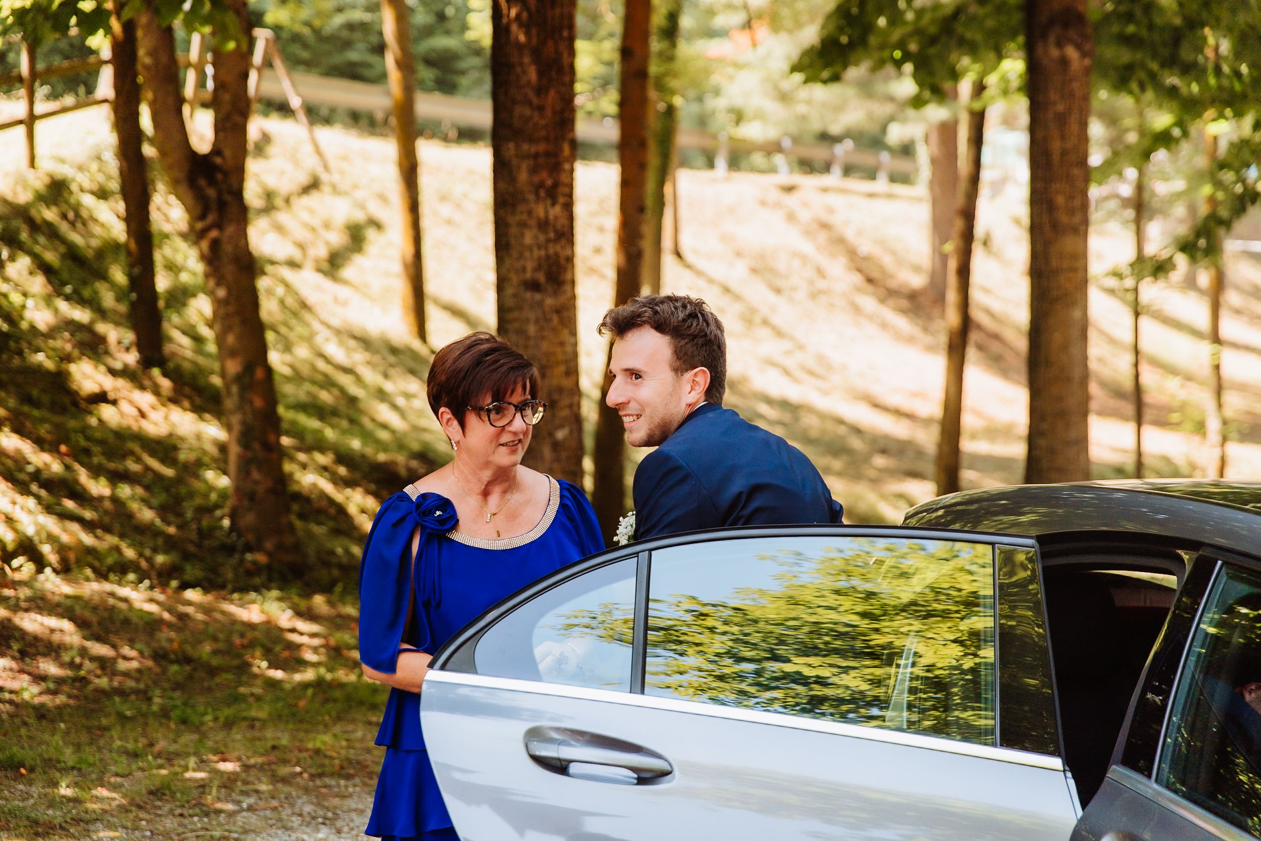 Couple enjoying a romantic moment in a sunny forest with a black car in the background.