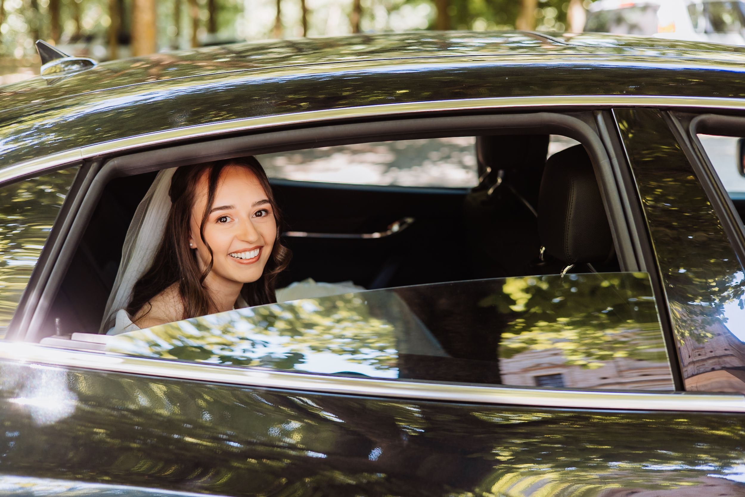 A woman looks through the window while sitting in a car with green foliage in the background.