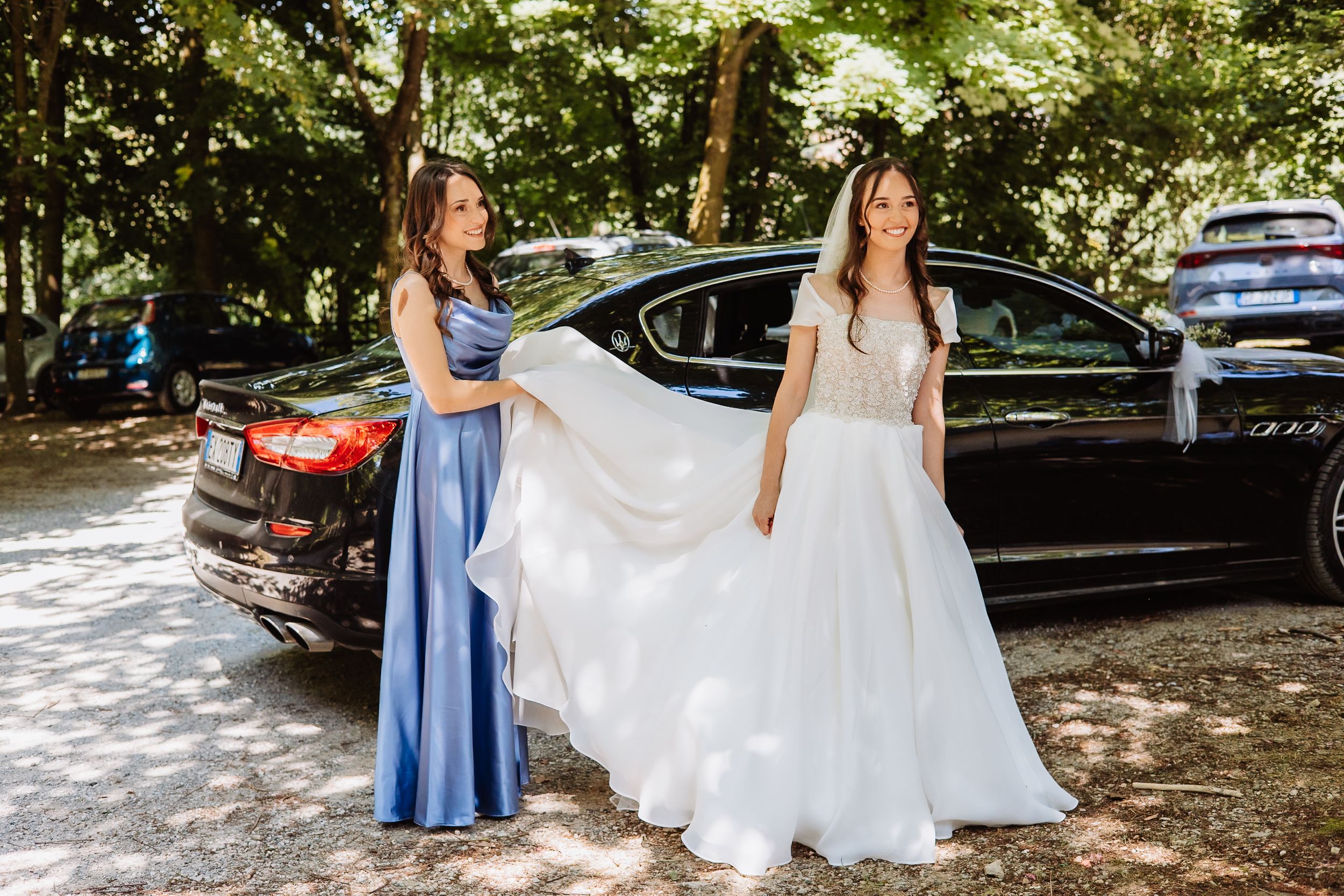 People in formal attire and flowing white dresses walking through a shaded outdoor area for a special event.