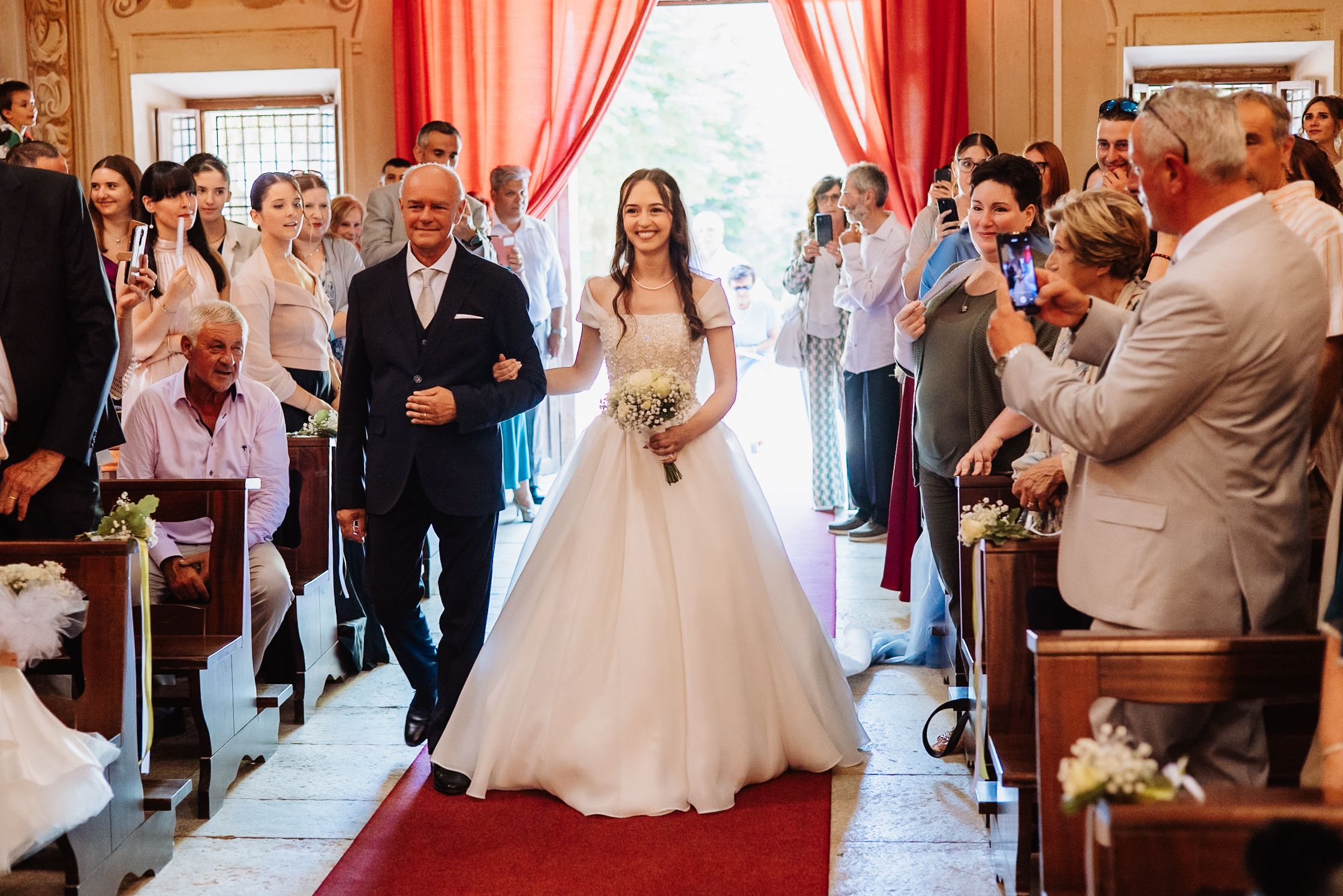 Wedding reception guests gather in an elegant room with red curtains and large windows.