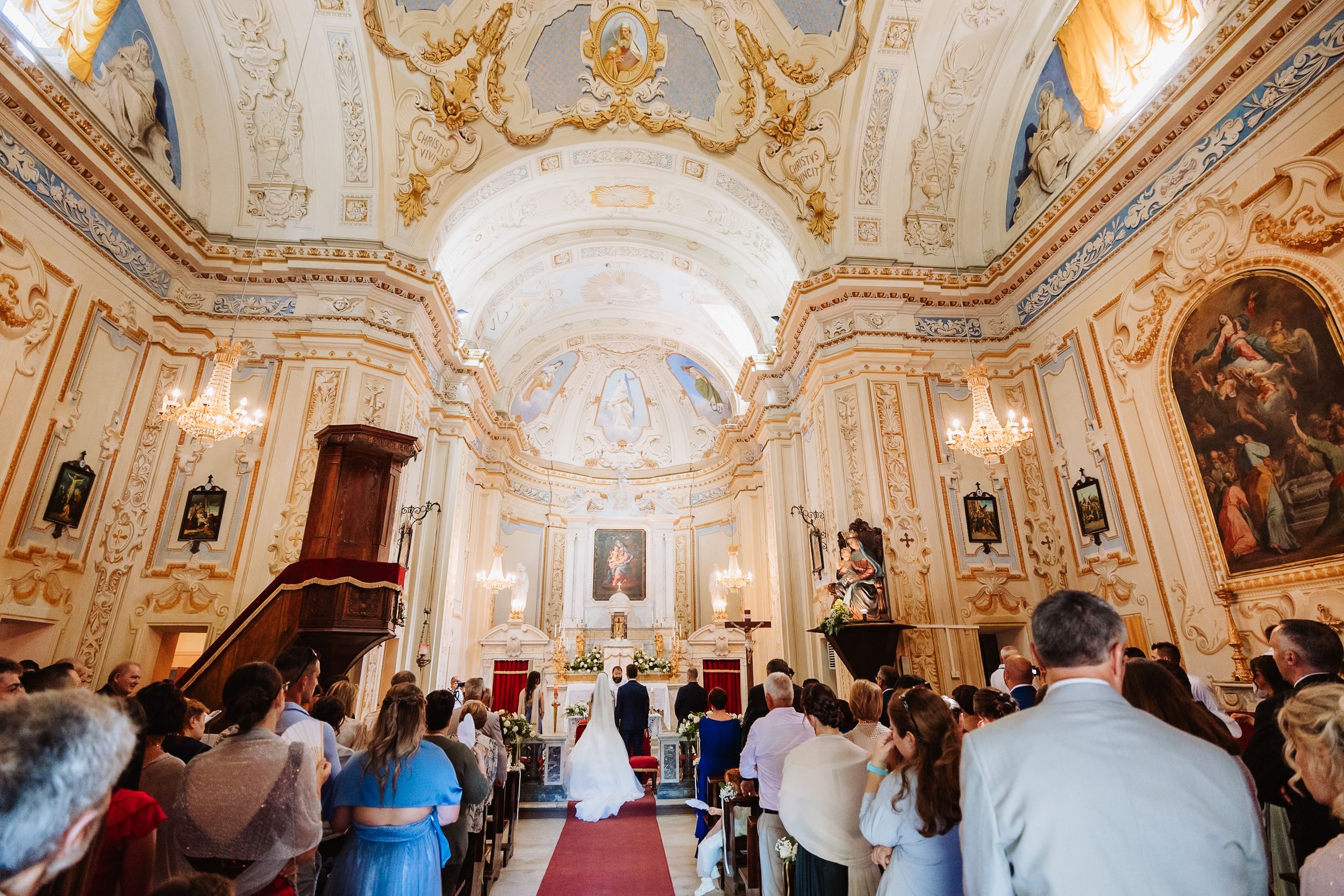 Visitors admire ornate Baroque architecture and artwork inside an opulent European palace with gilded decorations and frescoes.