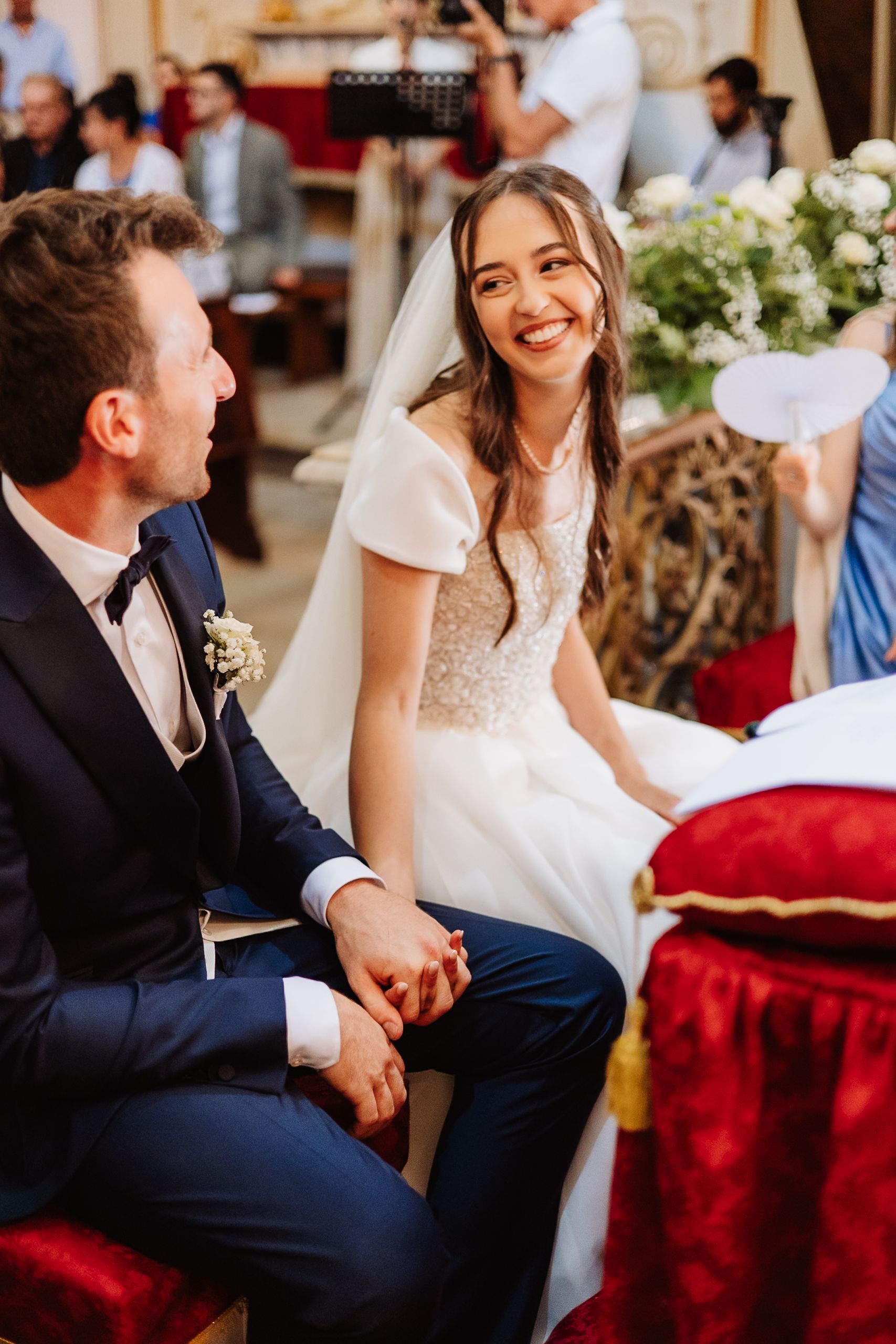 A joyful bride in a white lace dress sits beside her groom in a navy suit during their wedding ceremony.