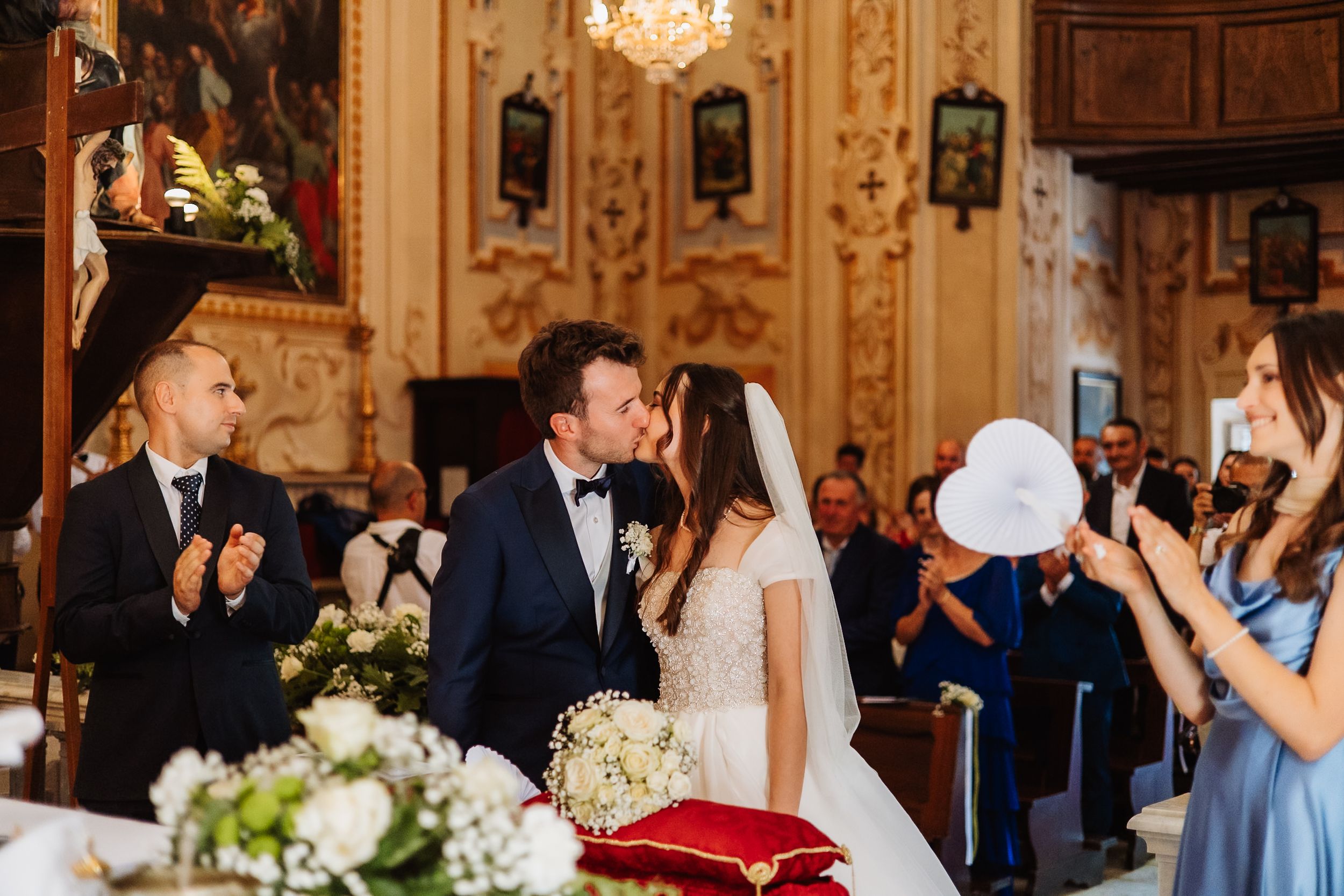 Wedding ceremony guests gather in an elegant ballroom with ornate decor, chandeliers and floral arrangements.