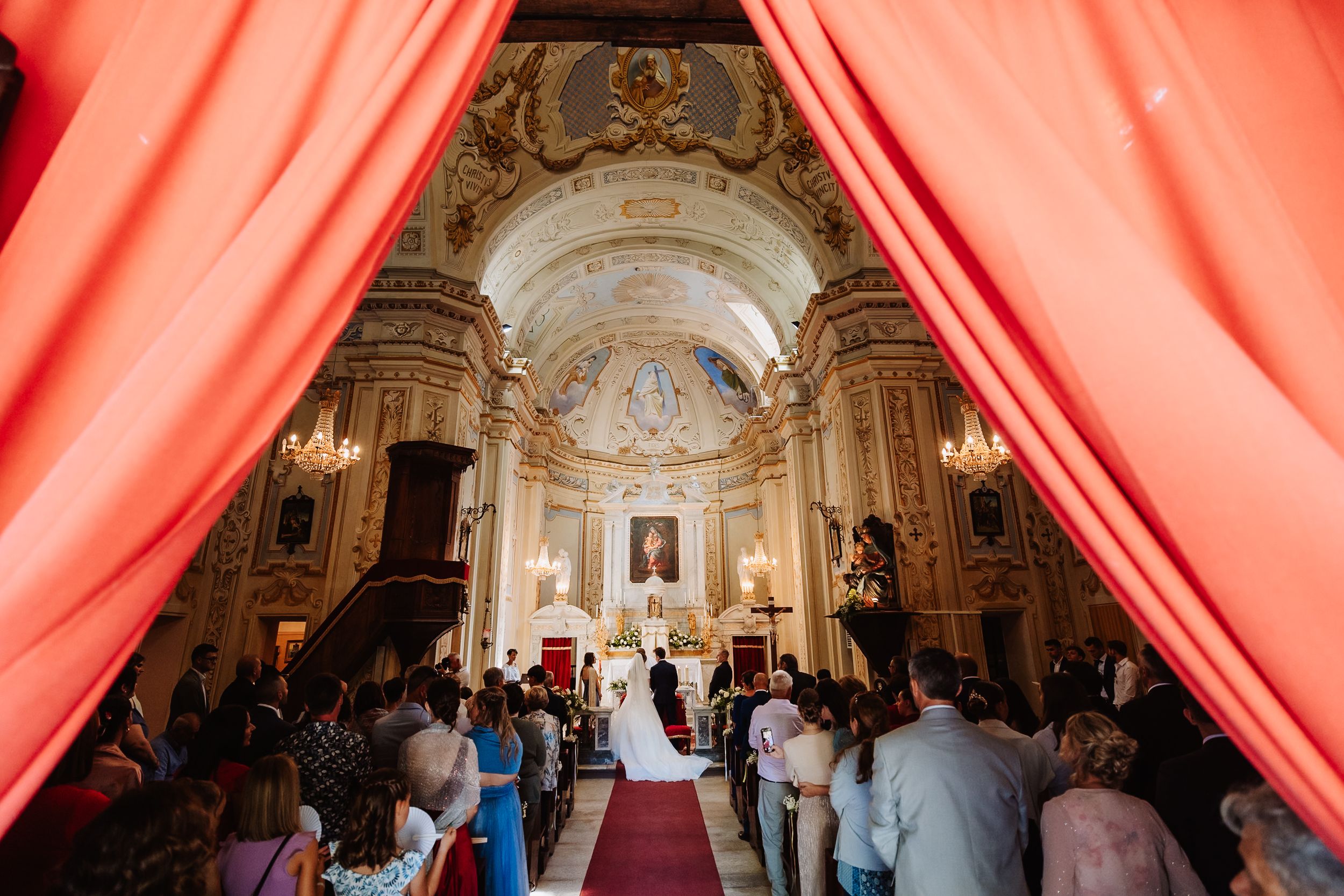 Wedding guests watch couple exchange vows during elegant ceremony in ornate cathedral with pink drapery decorations.