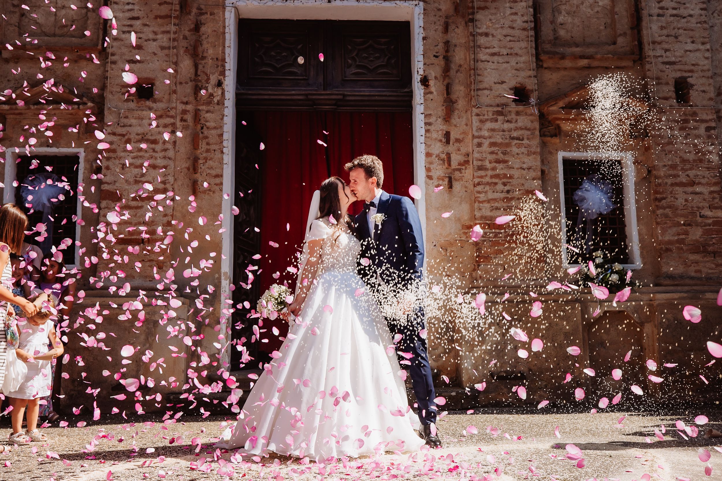 Newlywed couple exits historic brick church as guests shower them with pink and white flower petals in celebration.