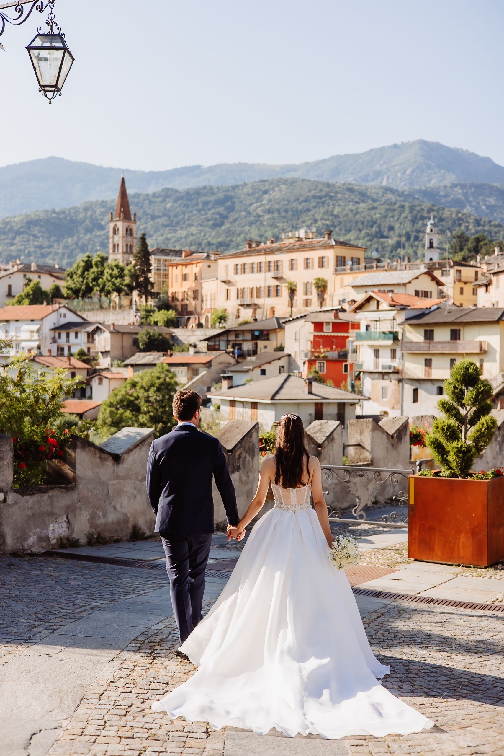 Wedding couple stands together overlooking historic Italian town with mountains and rooftops in panoramic view.