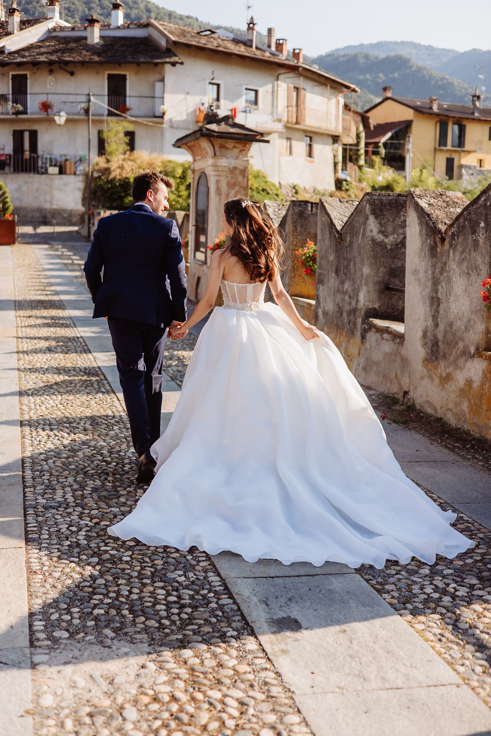 A couple walking along a cobblestone path in an Italian village, with the bride wearing a flowing white wedding gown.