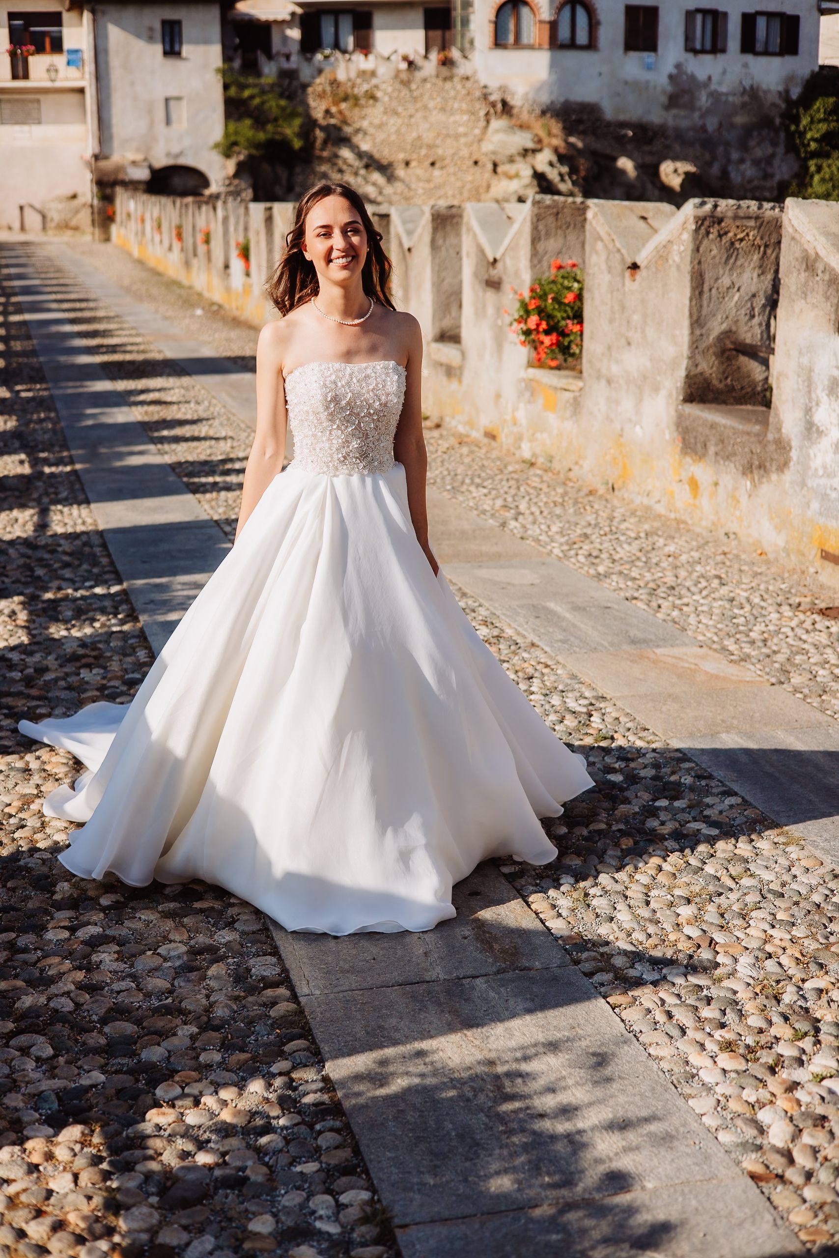 A wedding dress with a flowing white tulle skirt moves gracefully along a cobblestone pathway in sunlight.