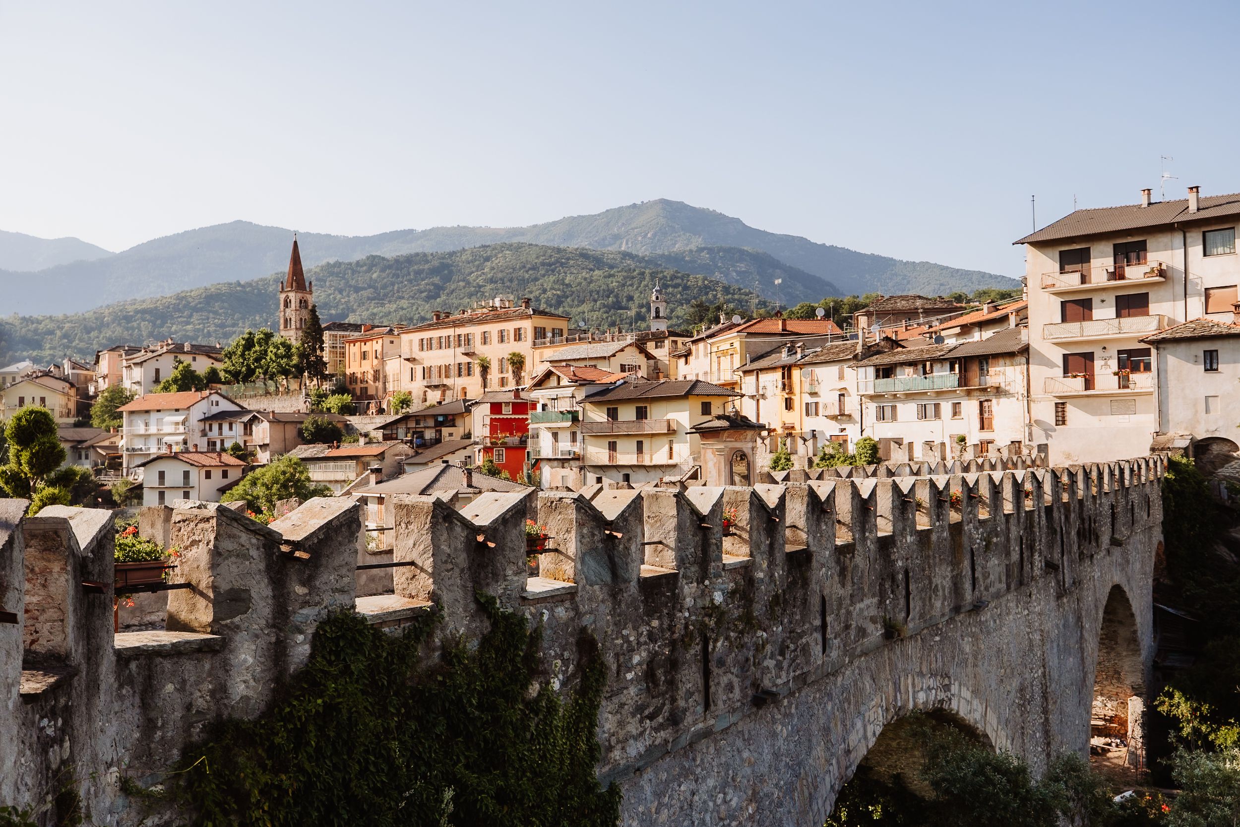 Ancient stone fortress wall overlooks a picturesque Italian hillside town with church spires and mountains in the background.