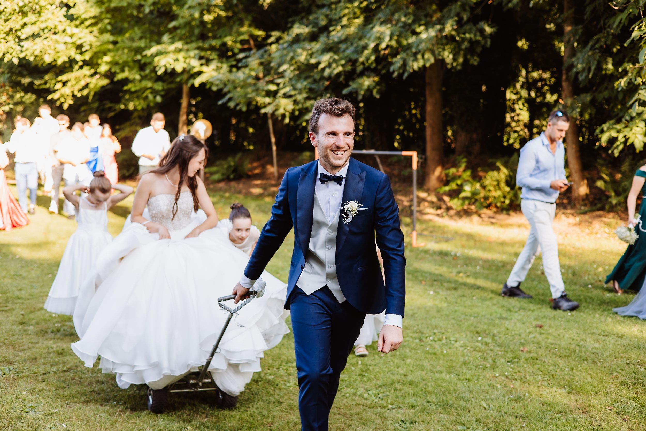 Wedding couple and guests gather in a garden setting for outdoor group photos during a sunny celebration.
