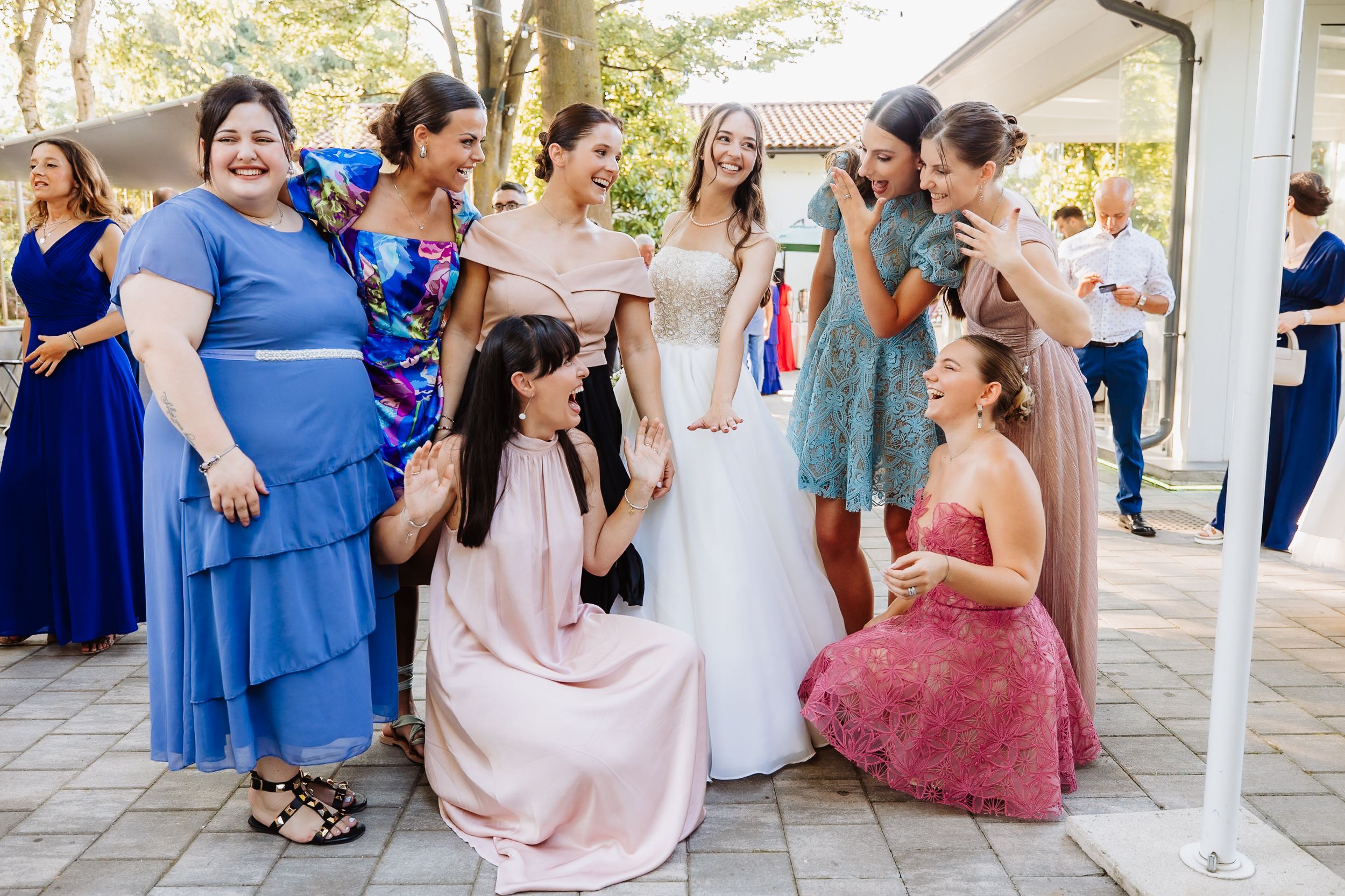A group in formal dresses gathers outdoors for a celebratory wedding photo at a venue patio.