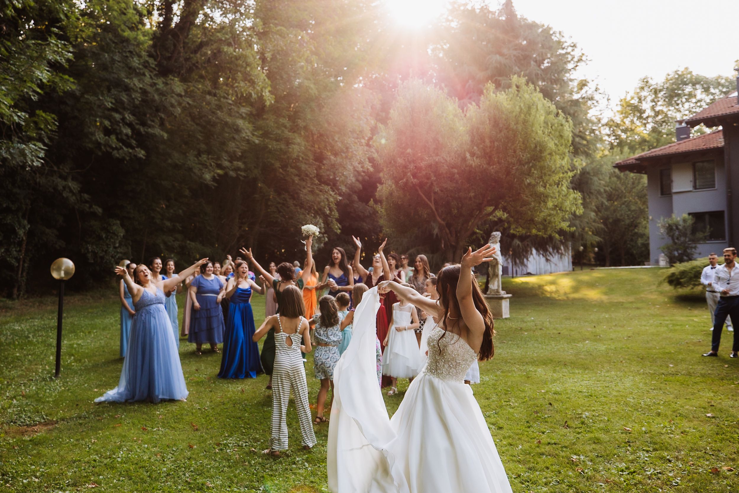 A row of guests stand in line outdoors at a sunny wedding reception surrounded by trees at golden hour.