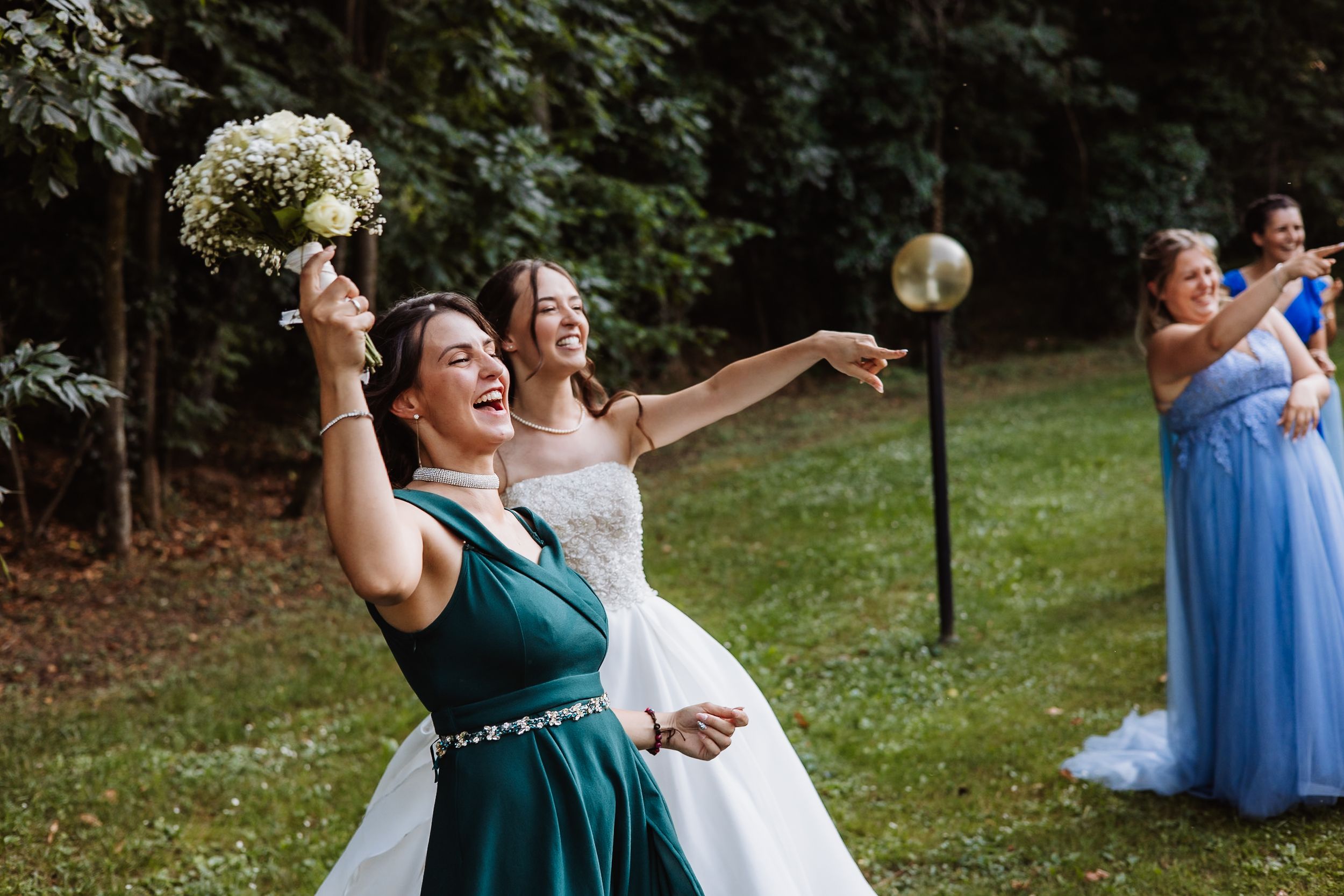 Wedding party in emerald green and light blue dresses laughing together outdoors in joyful celebration.