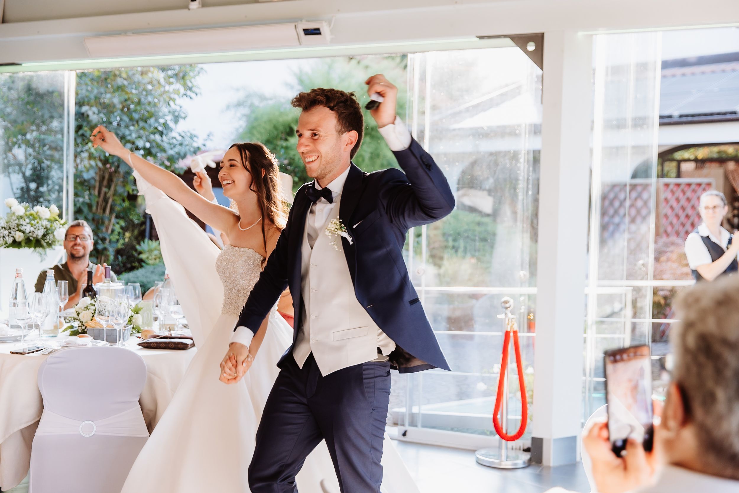A couple celebrates joyfully during their wedding reception in a bright, window-filled venue.