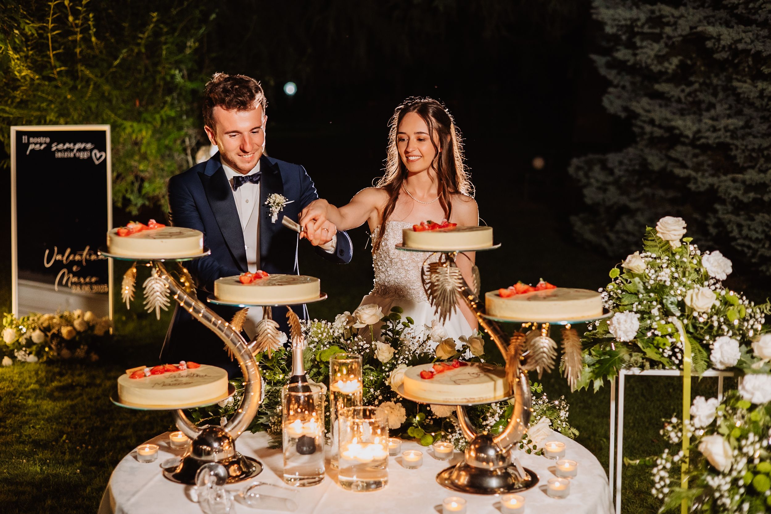 Wedding couple cuts into a rustic tiered cake at an elegant outdoor evening reception with glowing candle centerpieces.