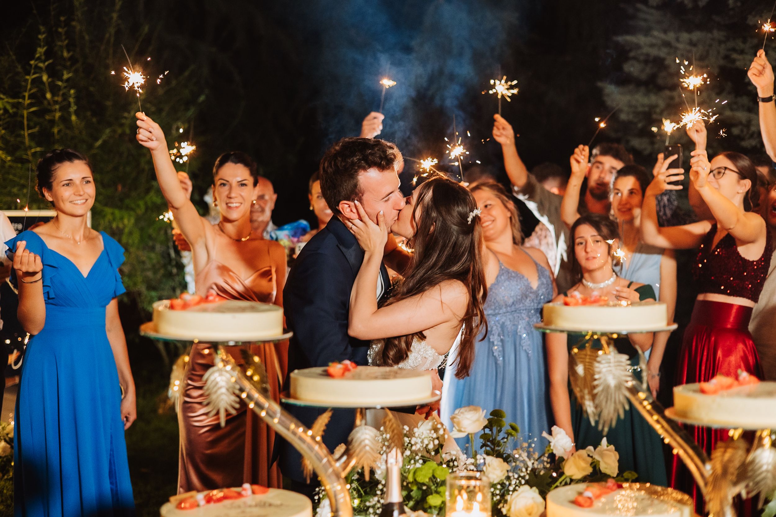 Guests hold sparklers and celebrate at an outdoor wedding reception in blue and black attire.
