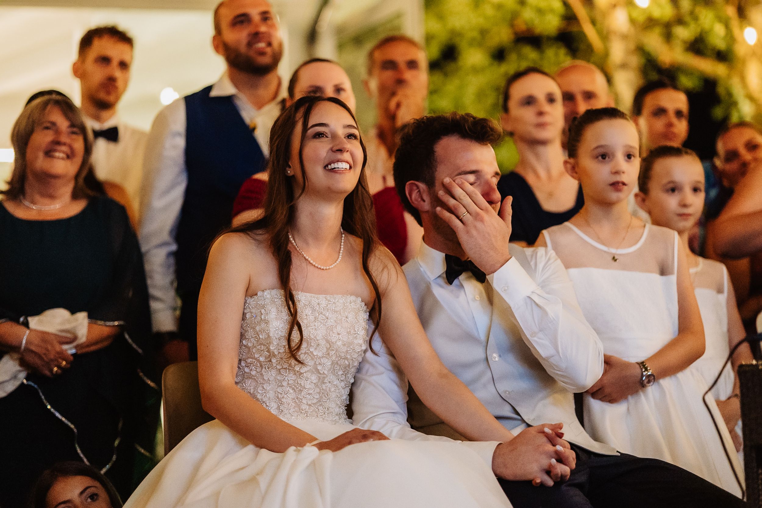 Wedding guests laugh and smile during an emotional reception moment in an outdoor setting.