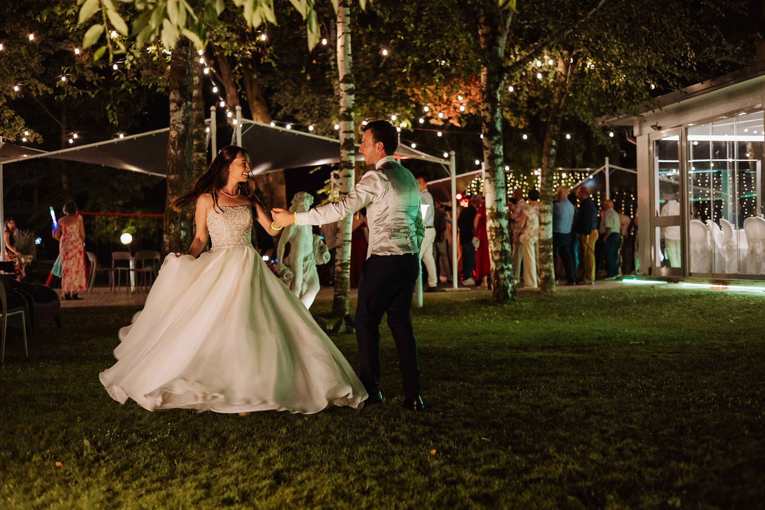 Wedding couple and guests dance together outdoors at sunset surrounded by string lights and trees.