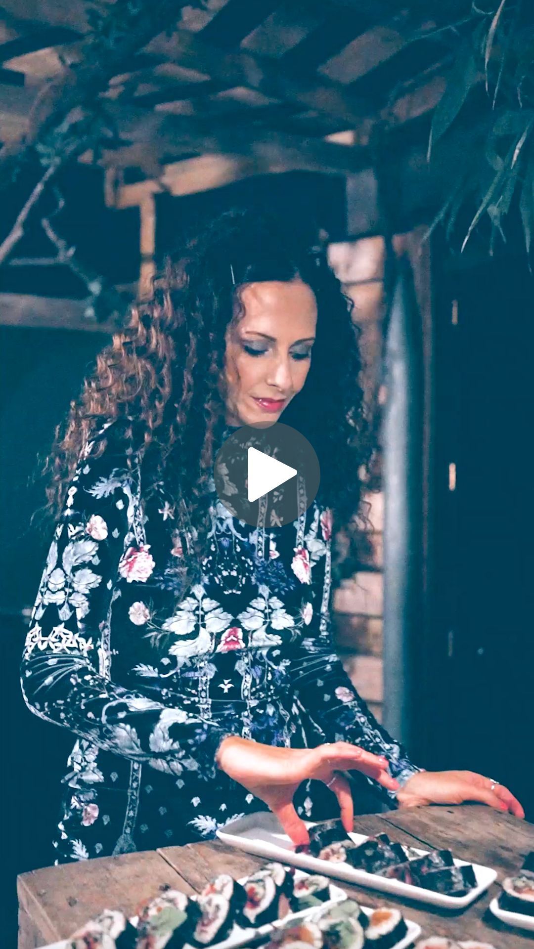 Person in floral dress prepares food at wooden counter in dimly lit space.