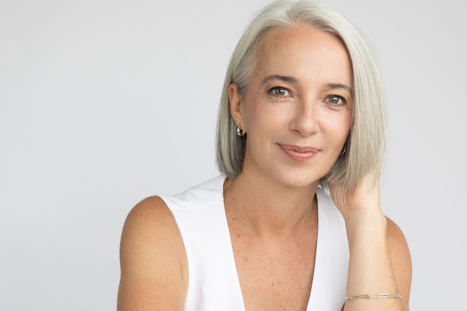 short silver haired woman with soft expression and hand on neck wearing a white vest with a gold bracelet on a white backdrop.