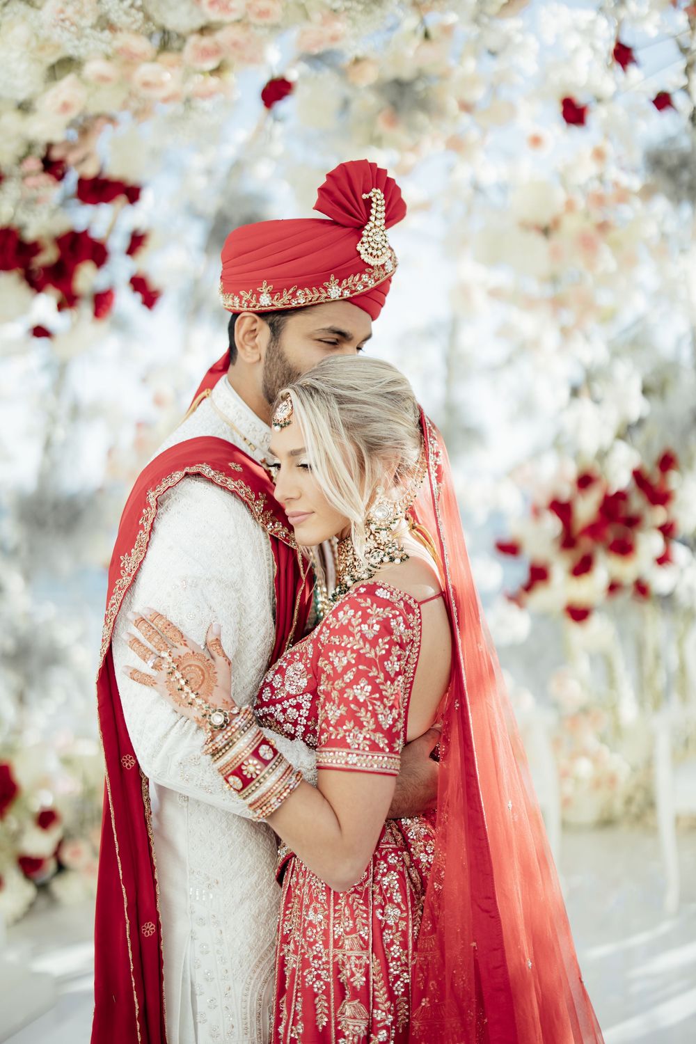 Traditional Indian wedding couple in red and white attire embracing amid falling rose petals.