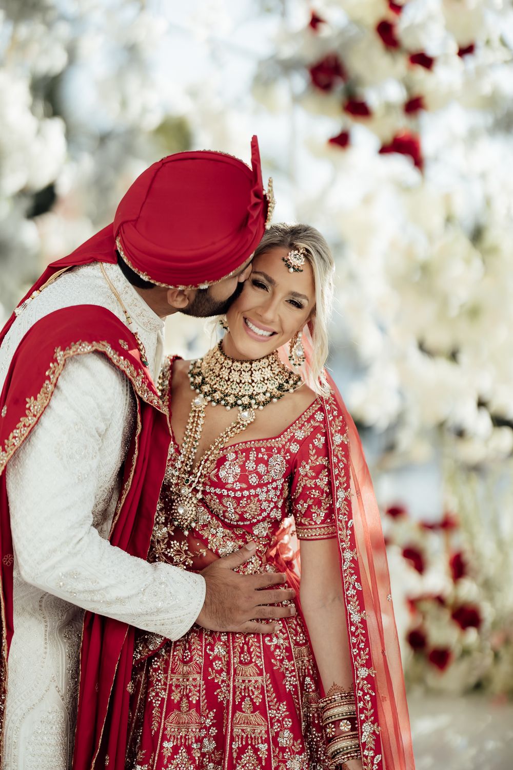 Traditional Indian wedding couple in red and white attire embrace under floral decor and white lights.