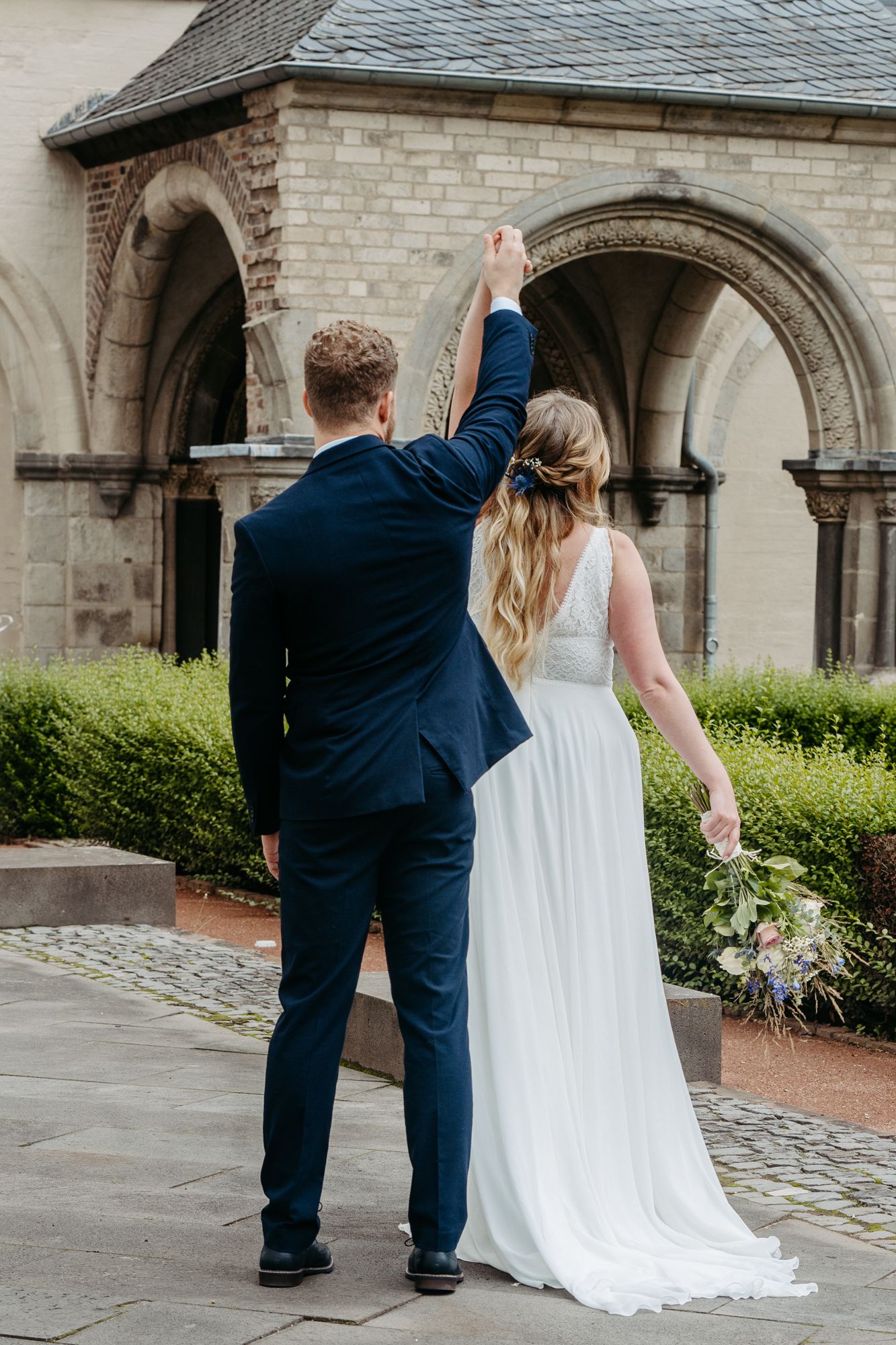 A wedding couple raises their arms in celebration outside a stone church archway.