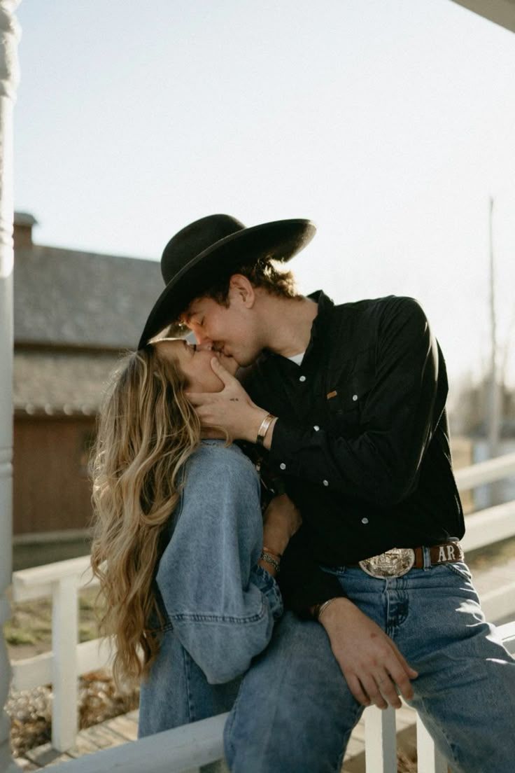 Romantic western-style couple in denim and black sharing an intimate moment on a rustic fence at sunset.
