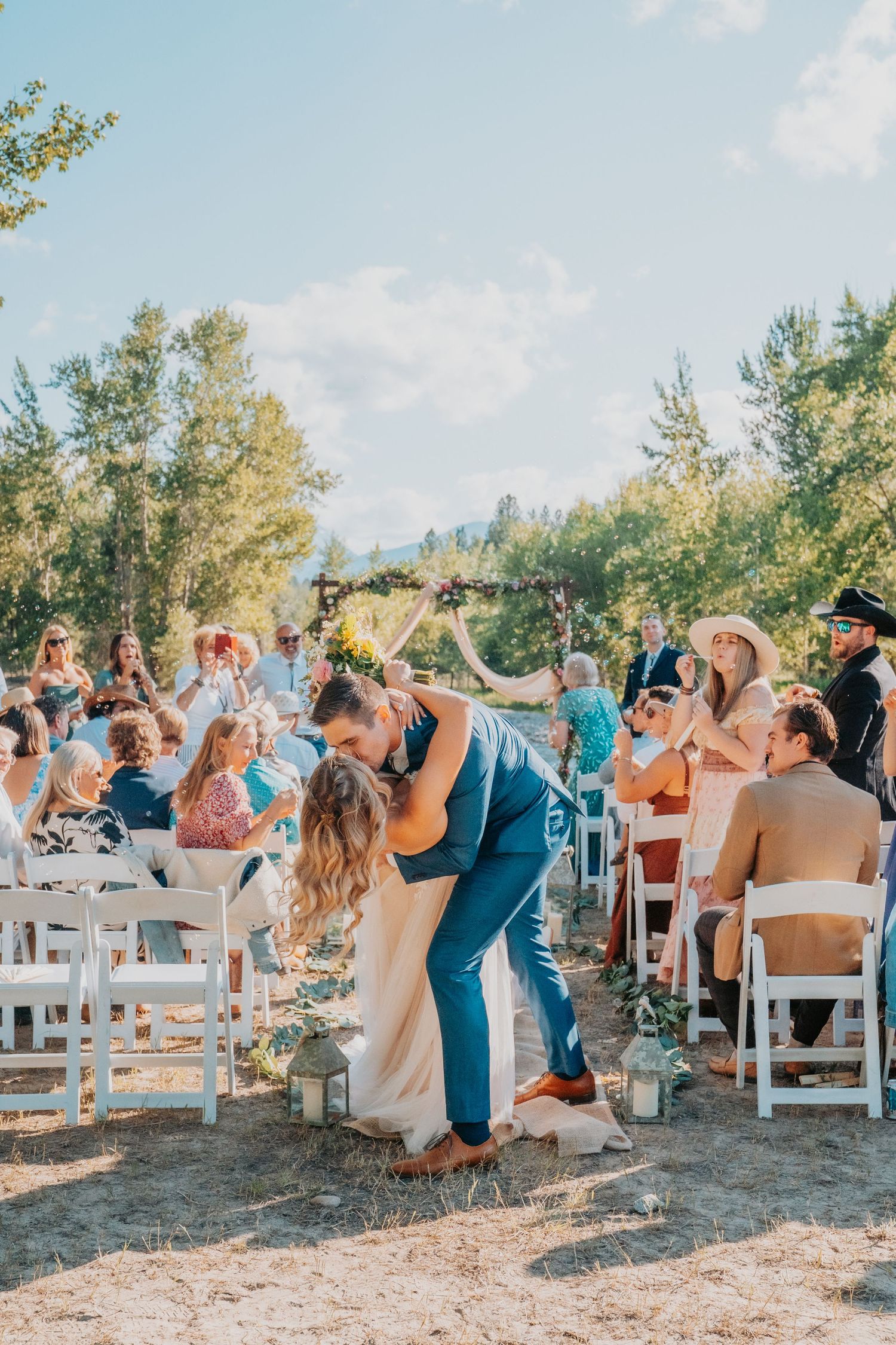 A joyful outdoor wedding ceremony with guests seated around a rustic wooden arch during golden hour.
