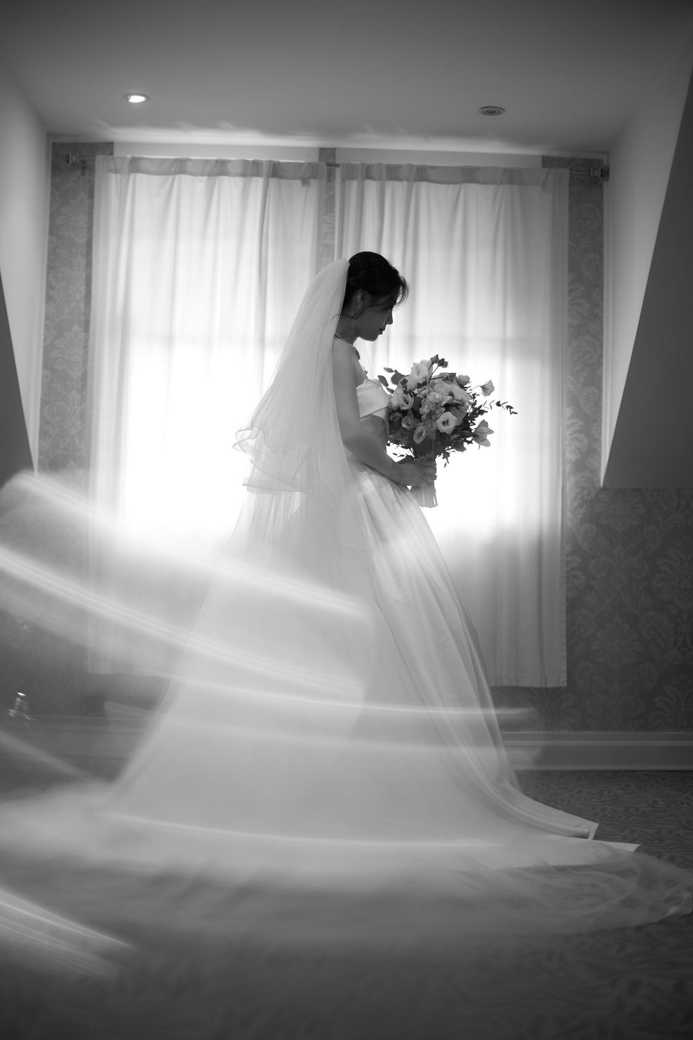A bride in silhouette stands by a window holding a bouquet, with her long veil flowing in black and white photography. at VIVO RESTAURANT
