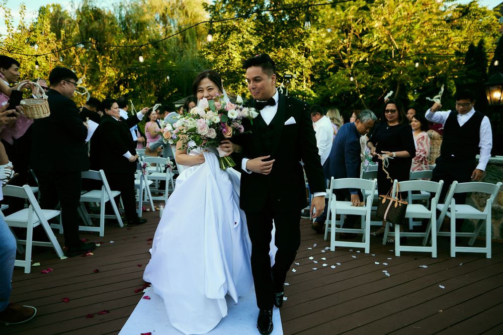 A wedding couple walks down the aisle together at an outdoor garden ceremony with white folding chairs. at VIVO RESTAURANT