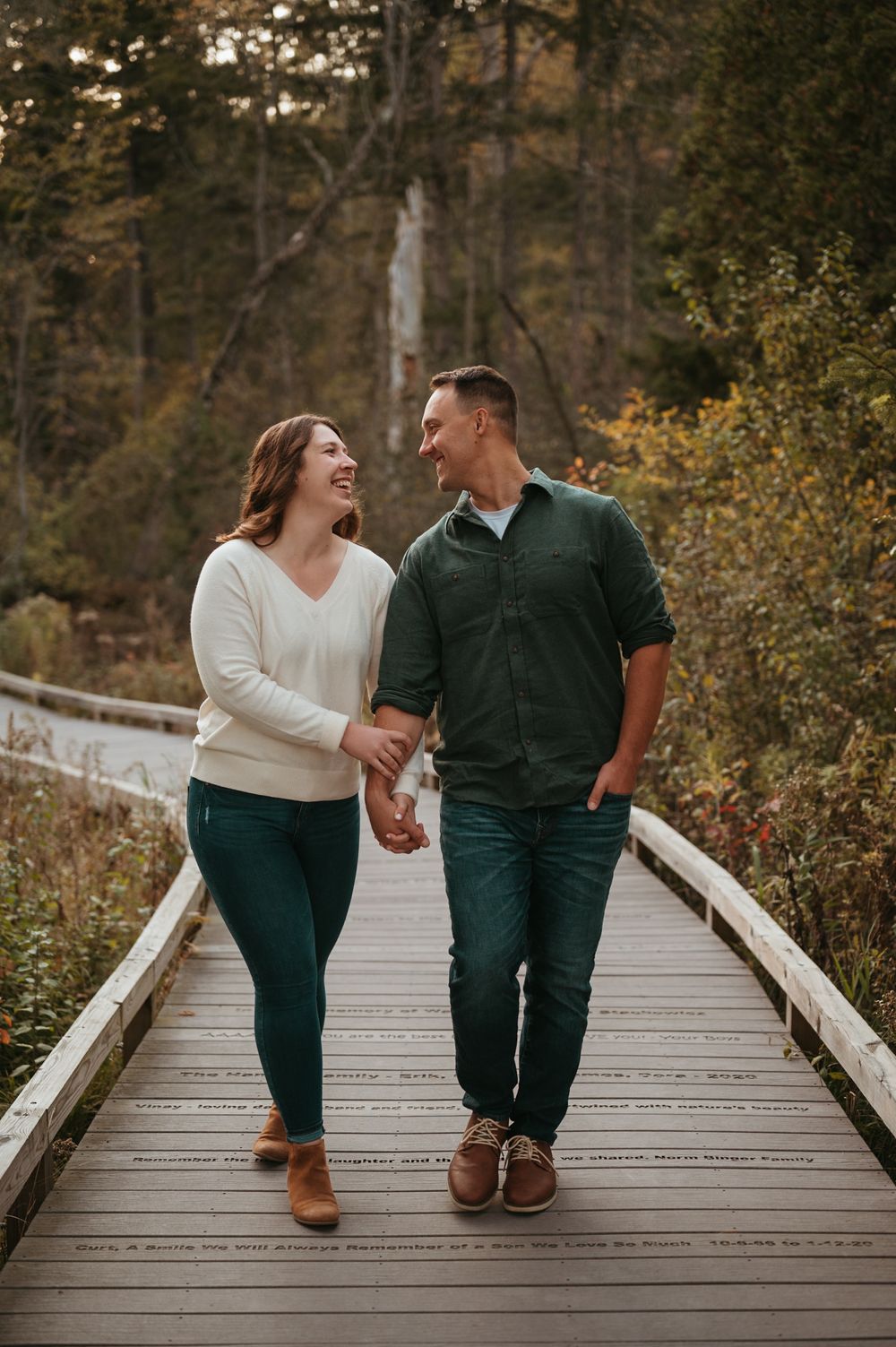 A couple laughing together while sitting on a wooden boardwalk surrounded by autumn foliage at sunset.