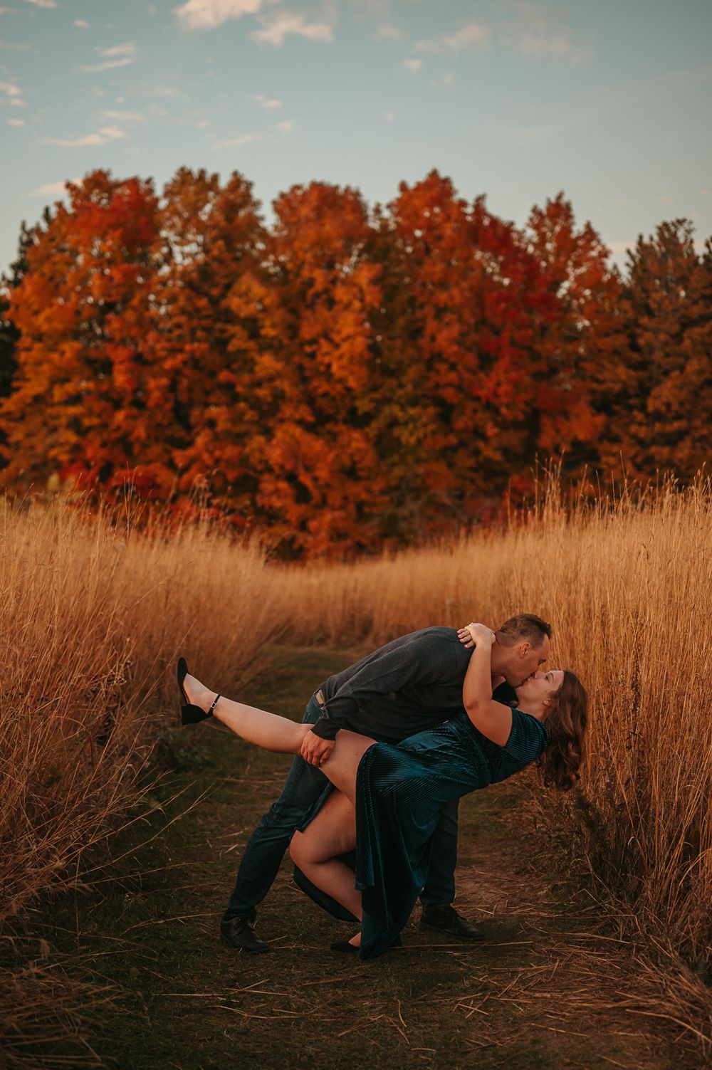 A romantic couple performs a dramatic dip pose in a field with vibrant orange autumn trees in the background.