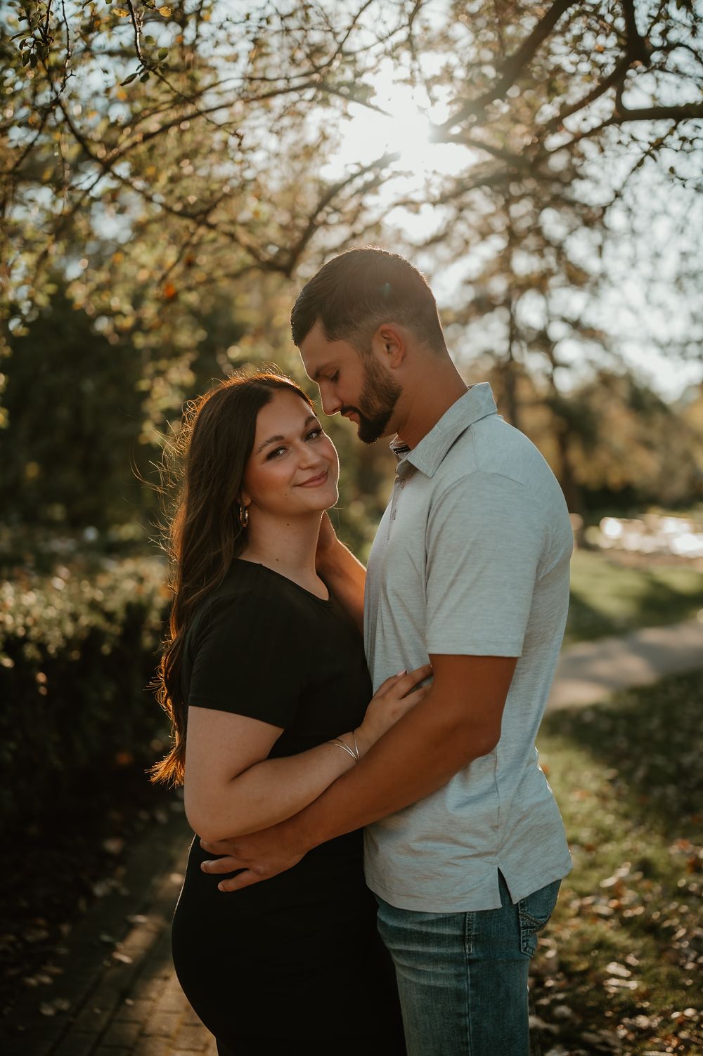 A couple embraces outdoors during golden hour with sunlit autumn trees in the background.