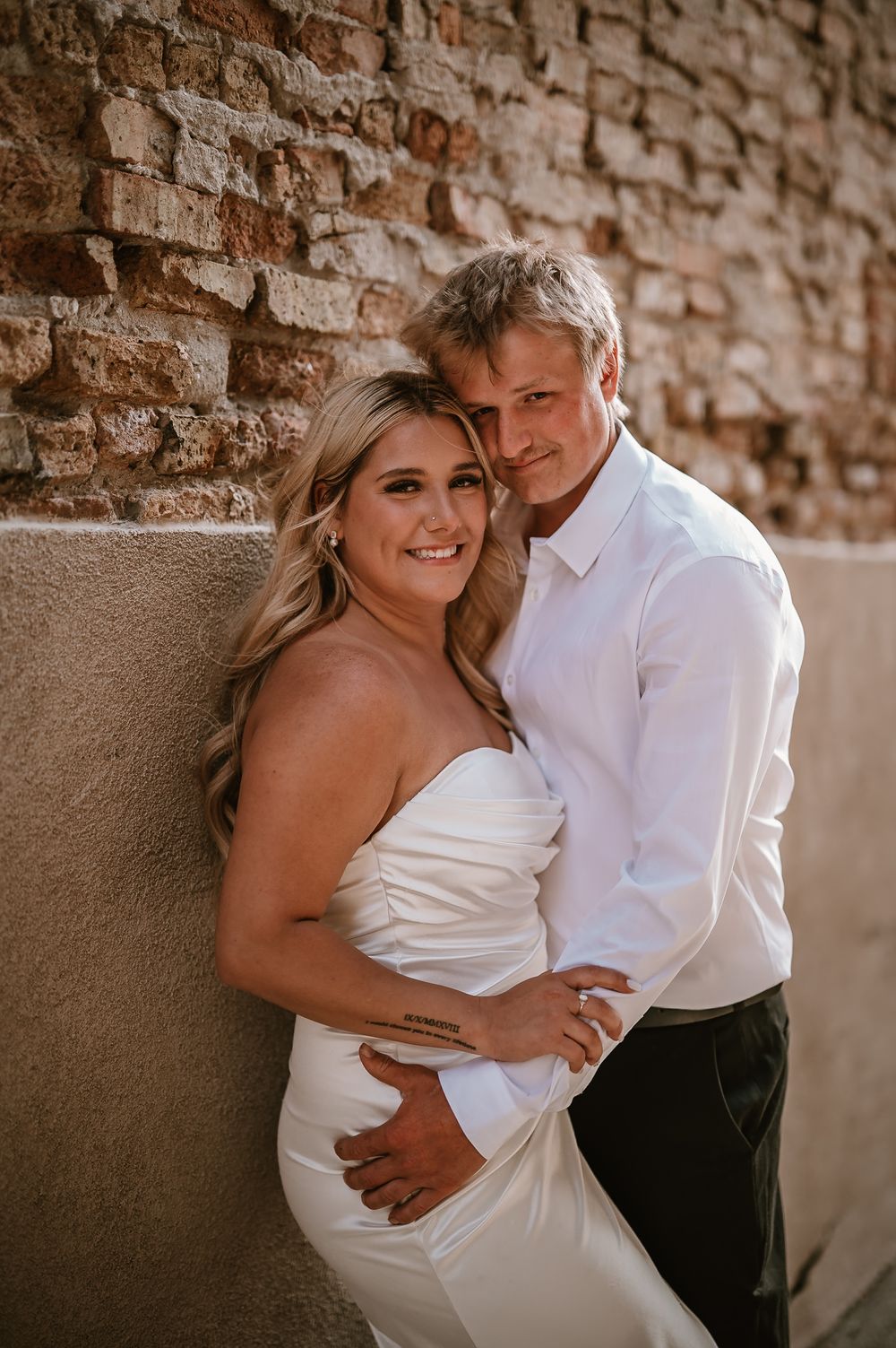 Romantic couple embraces against rustic brick wall as sun casts warm shadows during intimate photo session.