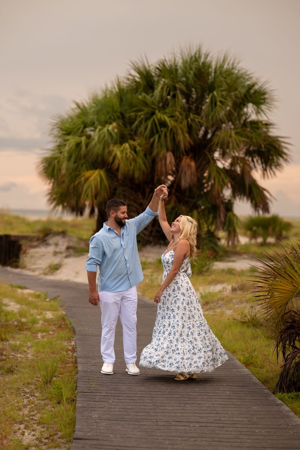A couple dancing together on a wooden boardwalk with palm trees and coastal scenery at sunset.