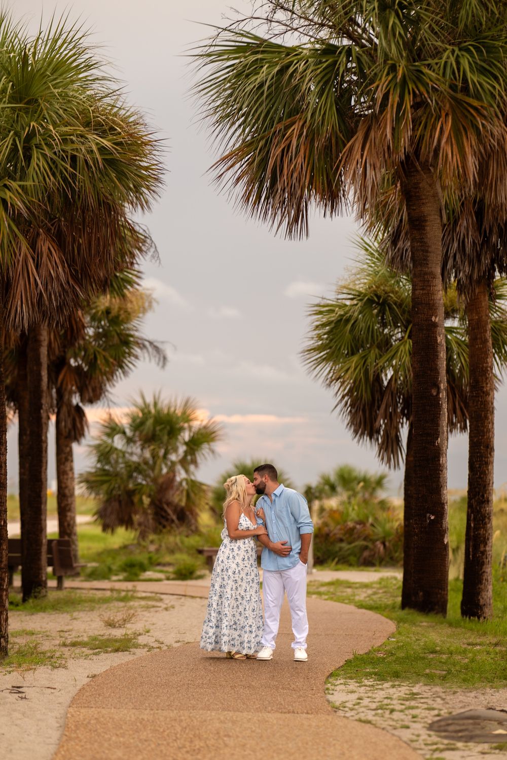 A couple walks along a palm tree lined path near the beach during golden hour.