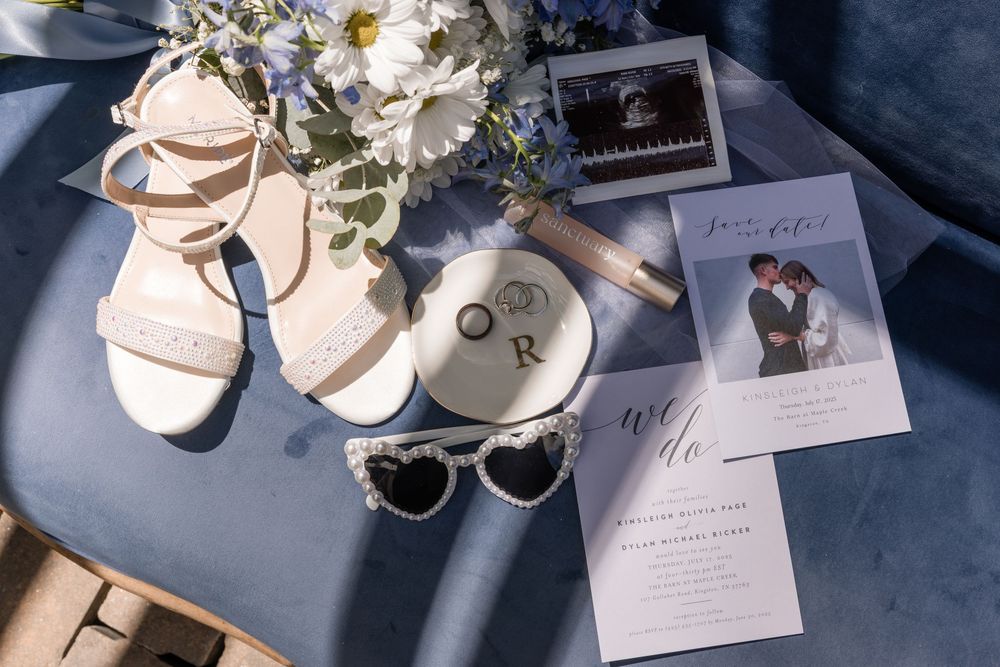 Wedding details flatlay with bridal shoes, sunglasses, invitation cards, rings and white floral bouquet on blue surface.