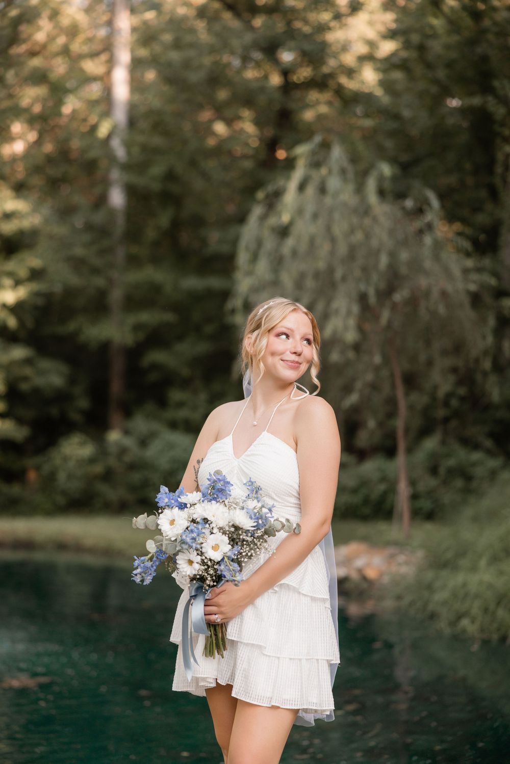 A bride in a short white dress holds a blue and white floral bouquet against a dark lake backdrop in the forest.